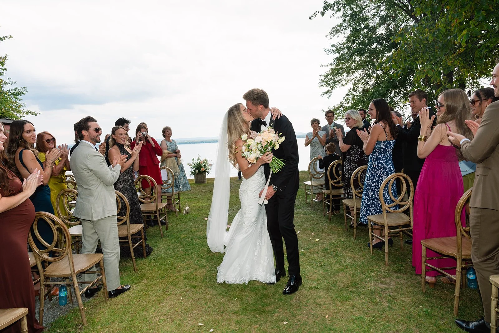 Bride and groom sharing a kiss at their outdoor wedding ceremony by a lake, surrounded by applauding guests.