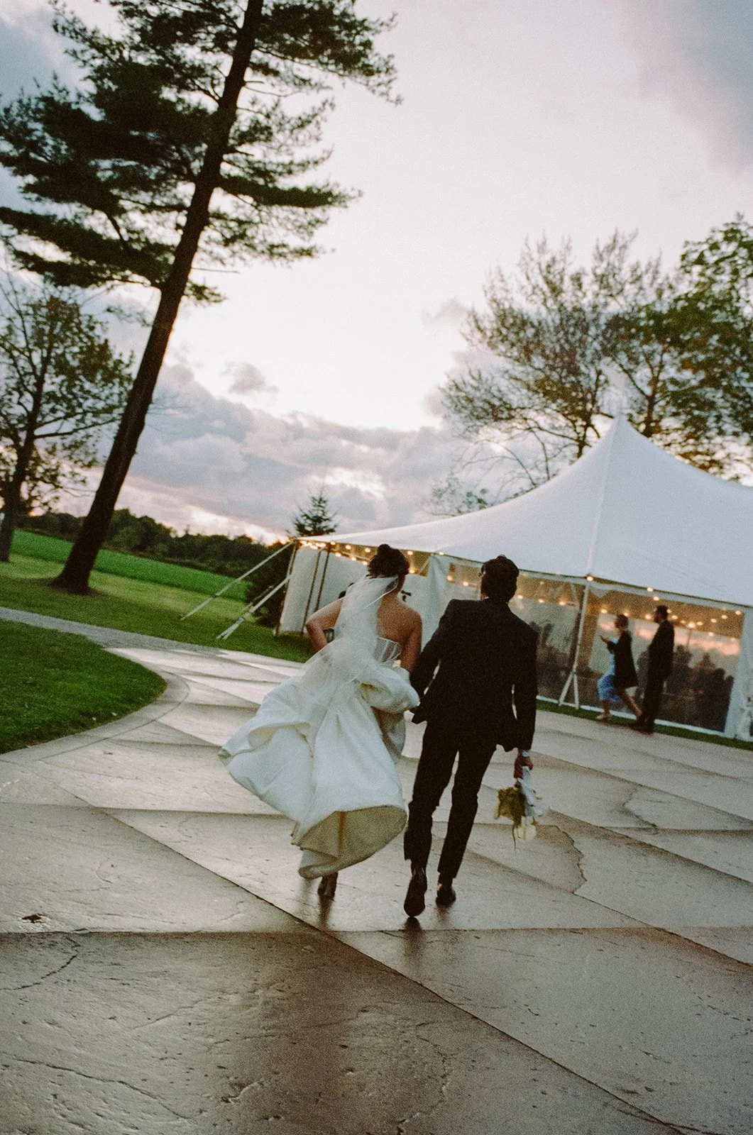 A bride and groom walking away from a wedding tent on a paved path during sunset.