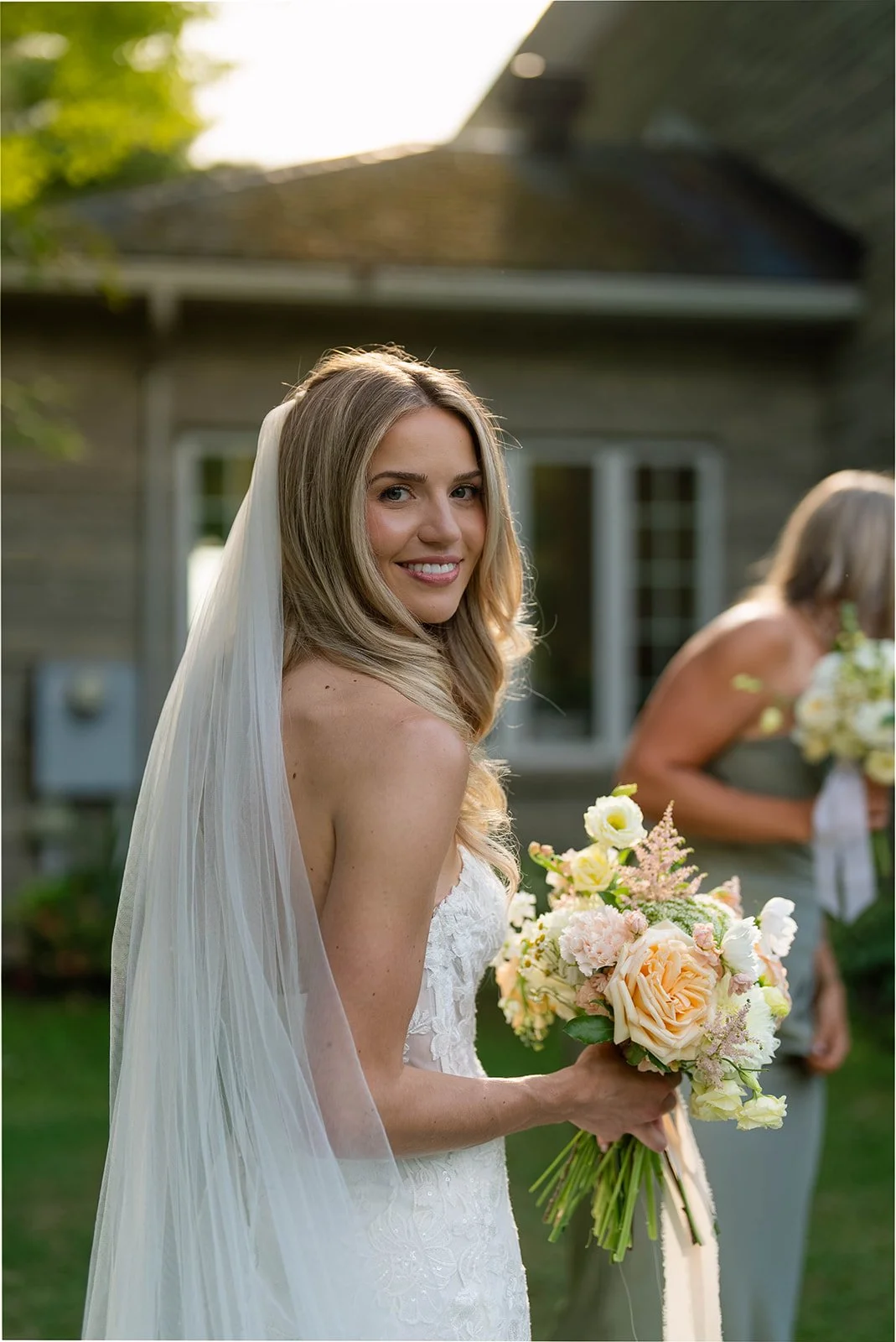 A bride with long blonde hair, wearing a white wedding dress with lace details and a veil, holding a bouquet of pastel-colored flowers, smiling outdoors during the daytime.