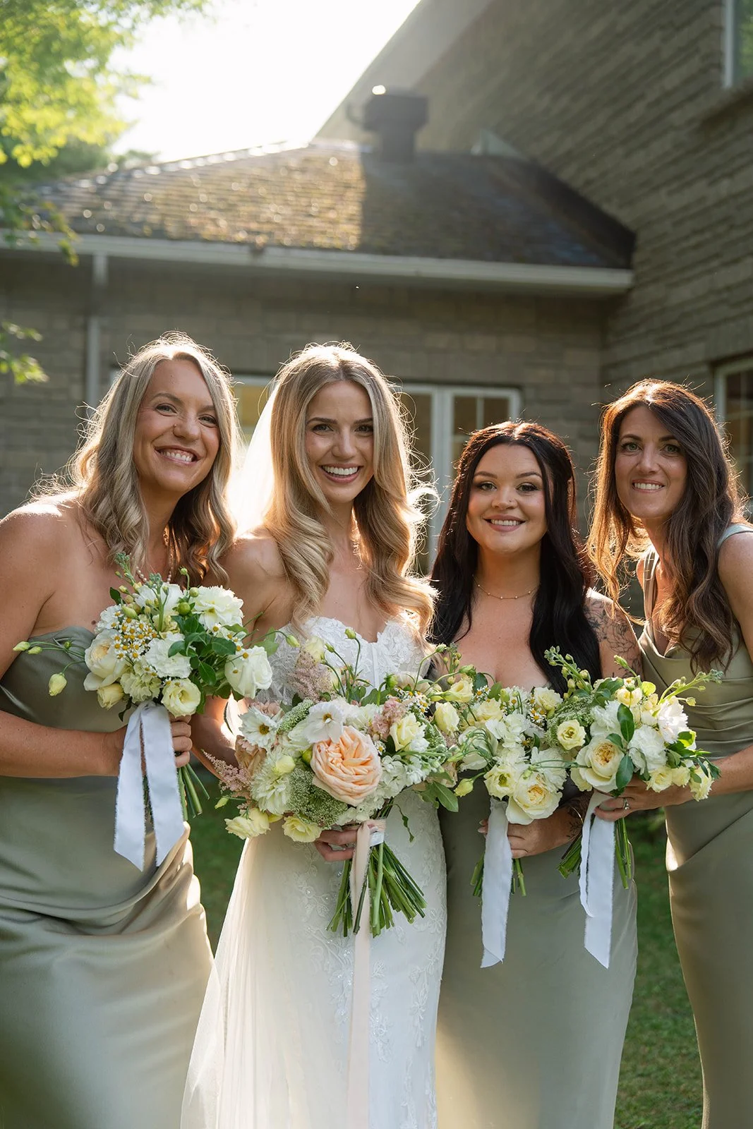 Four women dressed in formal attire standing outdoors, smiling, holding bouquets of flowers, with a building and trees in the background.