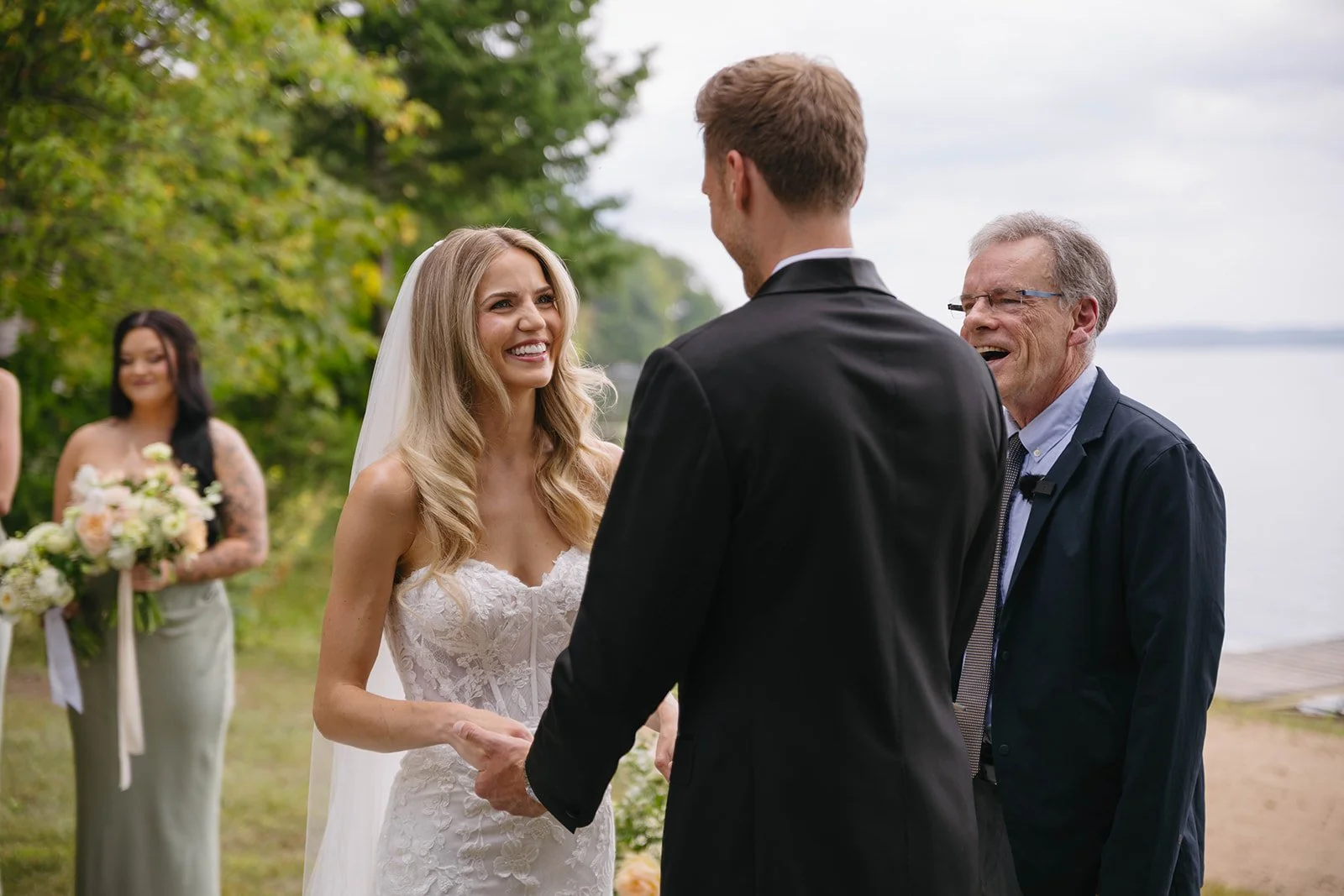 A bride and groom holding hands and smiling during their outdoor wedding ceremony by the water, with a woman holding a bouquet in the background.