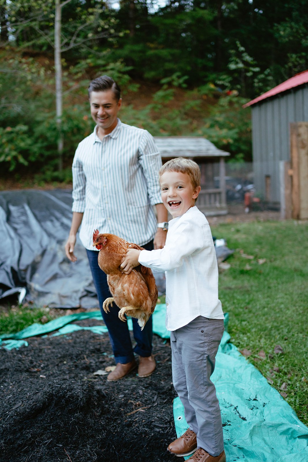 A young boy holding a chicken outdoors with a smiling woman standing behind him in a backyard.