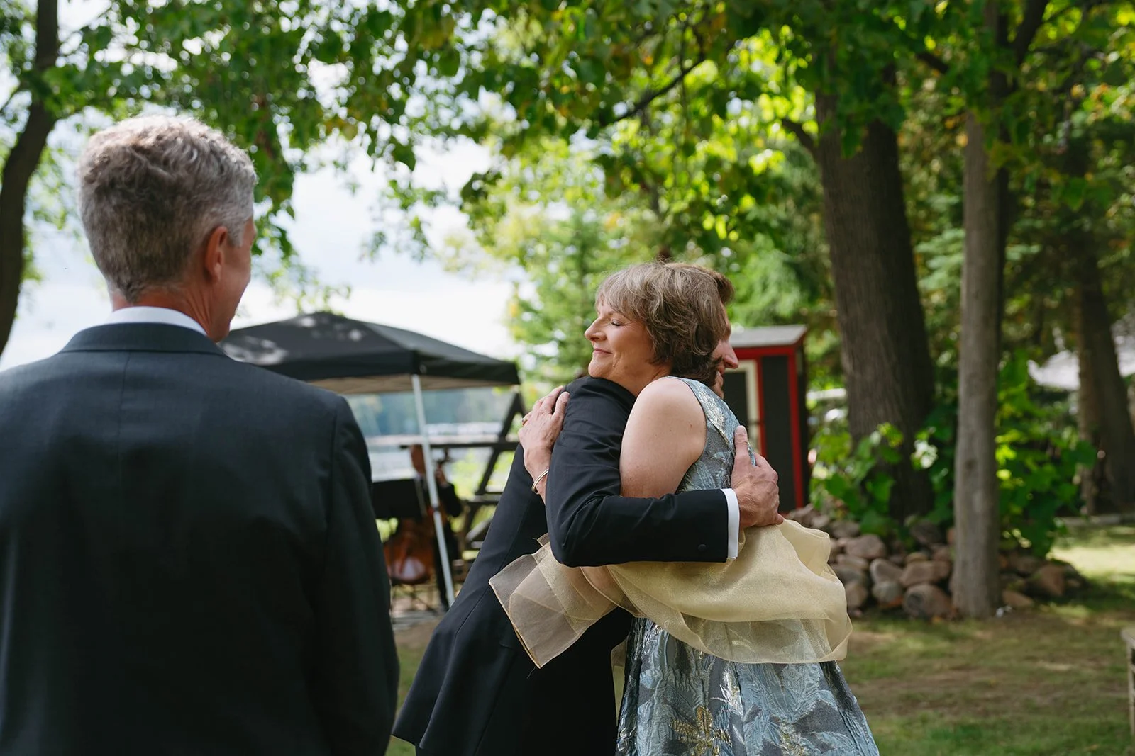 A woman hugging a man at outdoor event, with another man watching. They are surrounded by trees and outdoor furniture.
