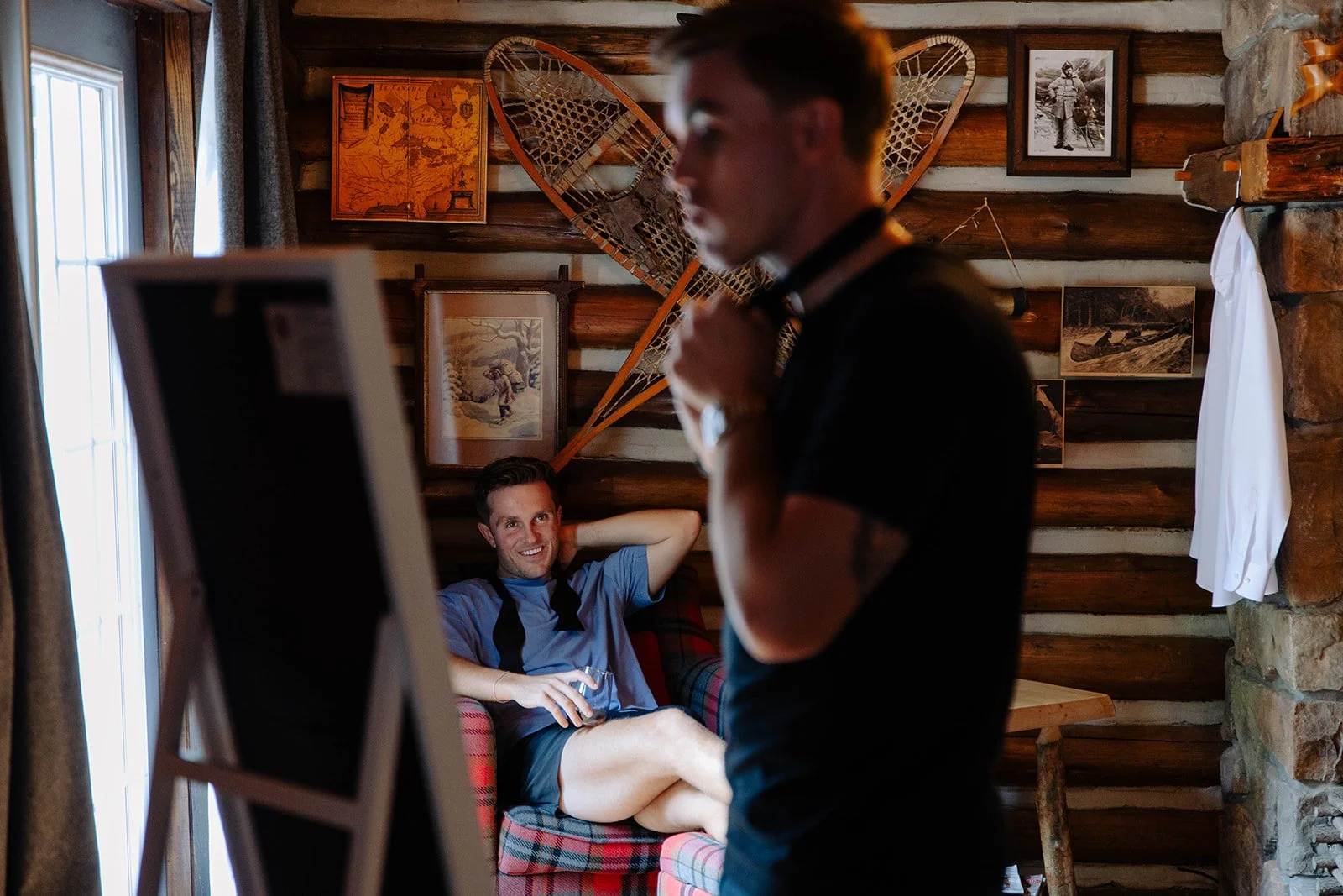 A young man sitting on a plaid armchair in a rustic cabin, smiling with a drink in hand, while another man adjusts a necktie or collar, with a wall decorated with vintage photos and snowshoes in the background.