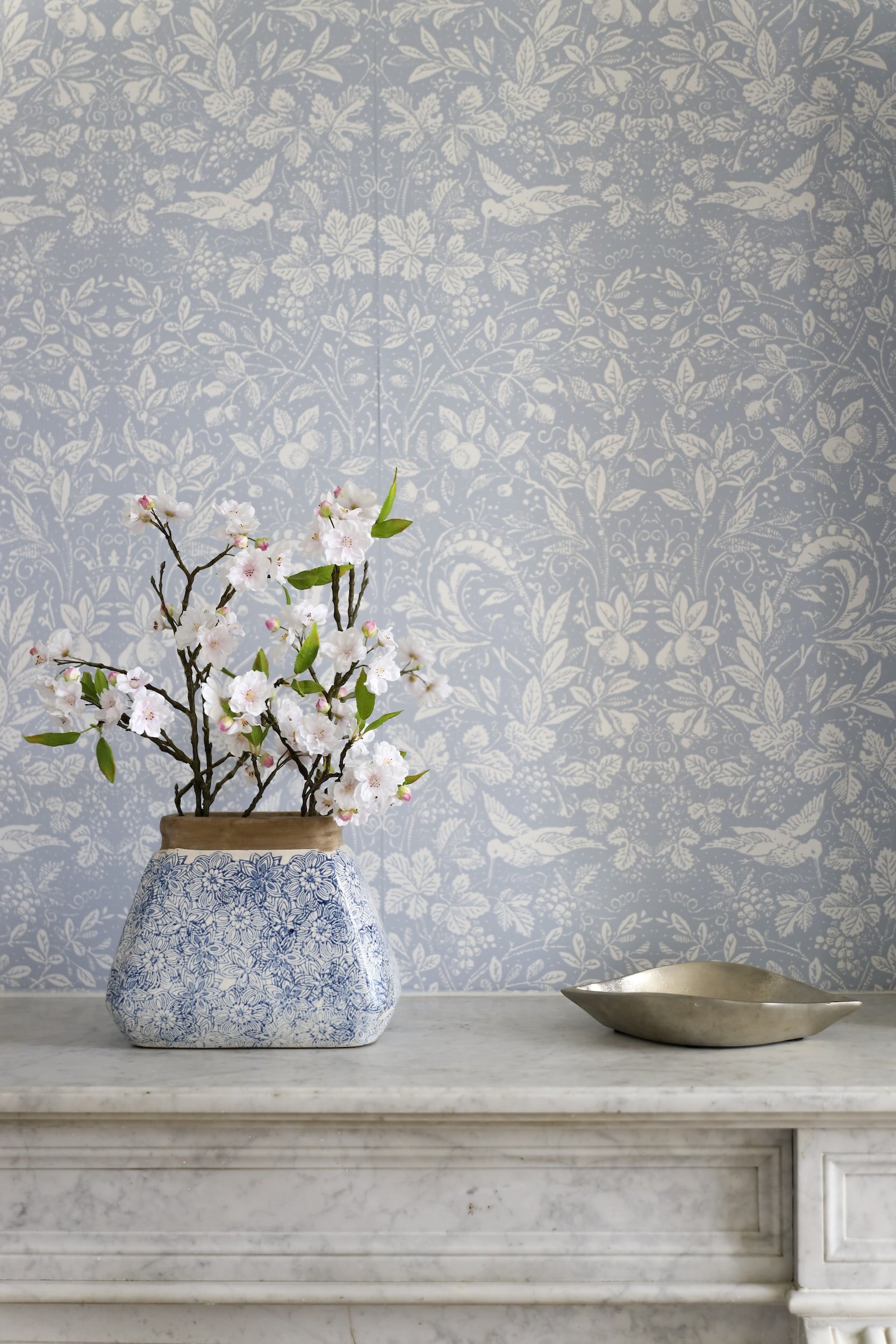 A floral arrangement of pink and white cherry blossoms in a blue and white patterned ceramic vase, placed on a white marble surface with a decorative blue wallpaper background.