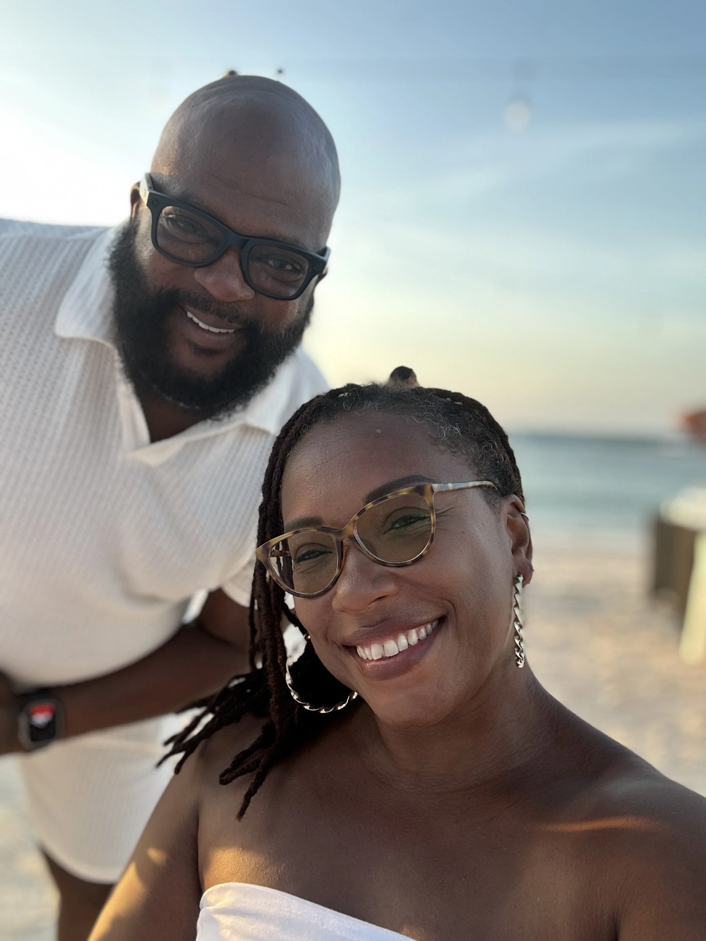 A smiling man and woman taking a selfie on the beach during sunset, with the ocean and sky in the background.