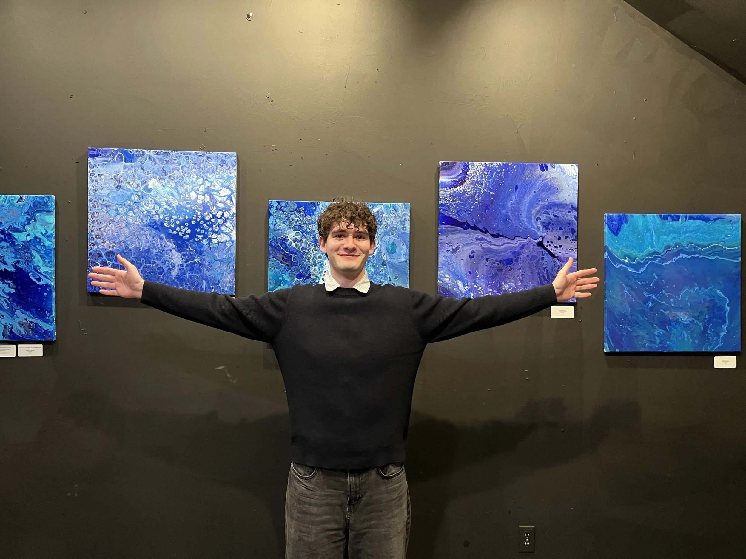 A young man with curly hair smiling and standing in front of a wall with five abstract blue paintings, arms outstretched.