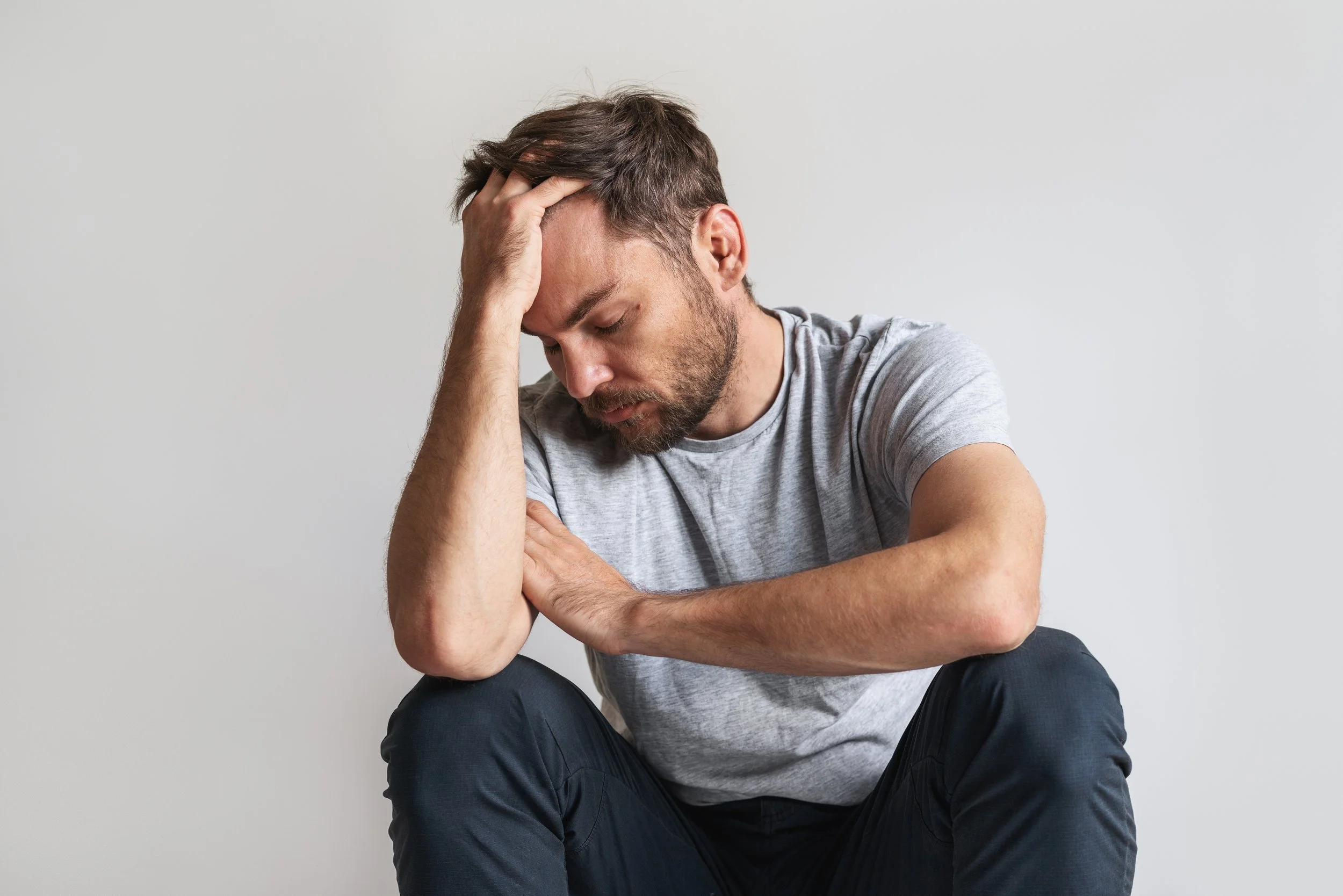 A photo of a man wearing dark blue pants and a light grey short-sleeved t-shirt sitting with his elbows on his knees, his head in his right hand, looking distressed.