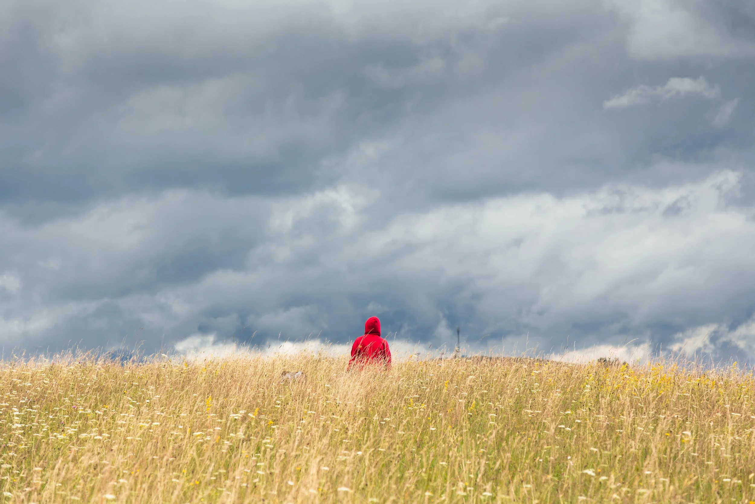A-lonely-man-inA person wearing a red hoodie stands alone on a grassy hill under a dramatic, cloudy sky in tranquility and solitude.