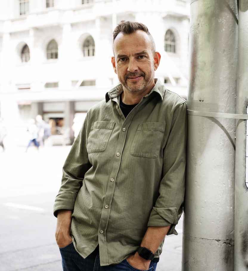 Jeremy Macvean wearing a green button-up shirt with rolled-up sleeves and a black watch, leaning against a large metallic pole on a city street.