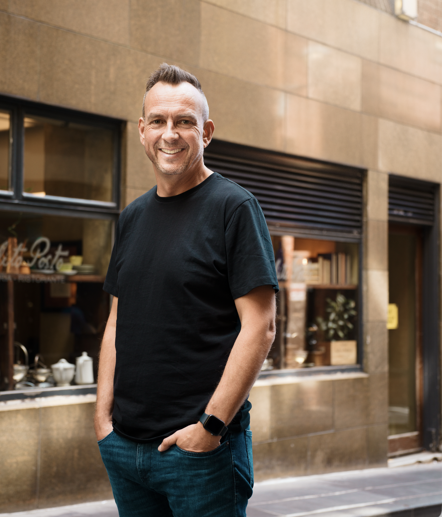 Jeremy Macvean wearing a black t-shirt and blue jeans, with a smartwatch on his left wrist, standing on a city street in front of a windowed storefront with shelves of dishes and decor inside.