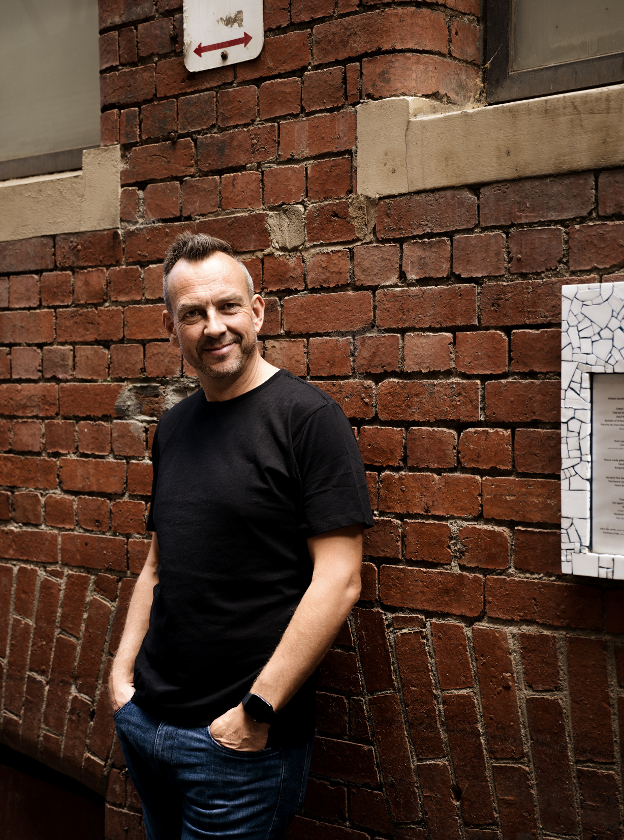 Jeremy Macvean wearing a black t-shirt and jeans, standing against a red brick wall with his hands in his pockets, smiling at the camera.