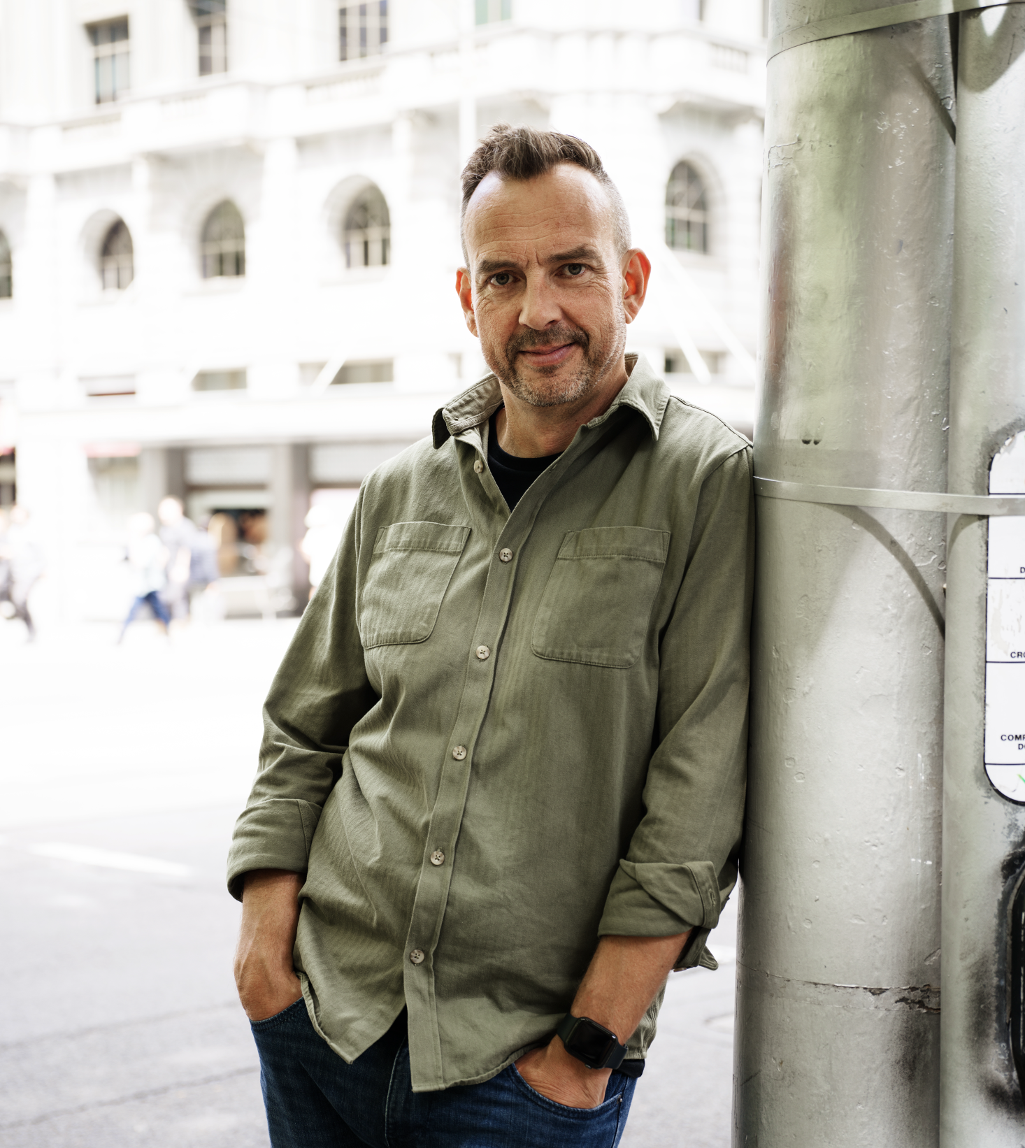 Jeremy Macvean leaning against a pole on a city street. He is wearing a green button-up shirt with rolled-up sleeves, dark jeans, and a smartwatch. The background shows blurred pedestrians and urban buildings.
