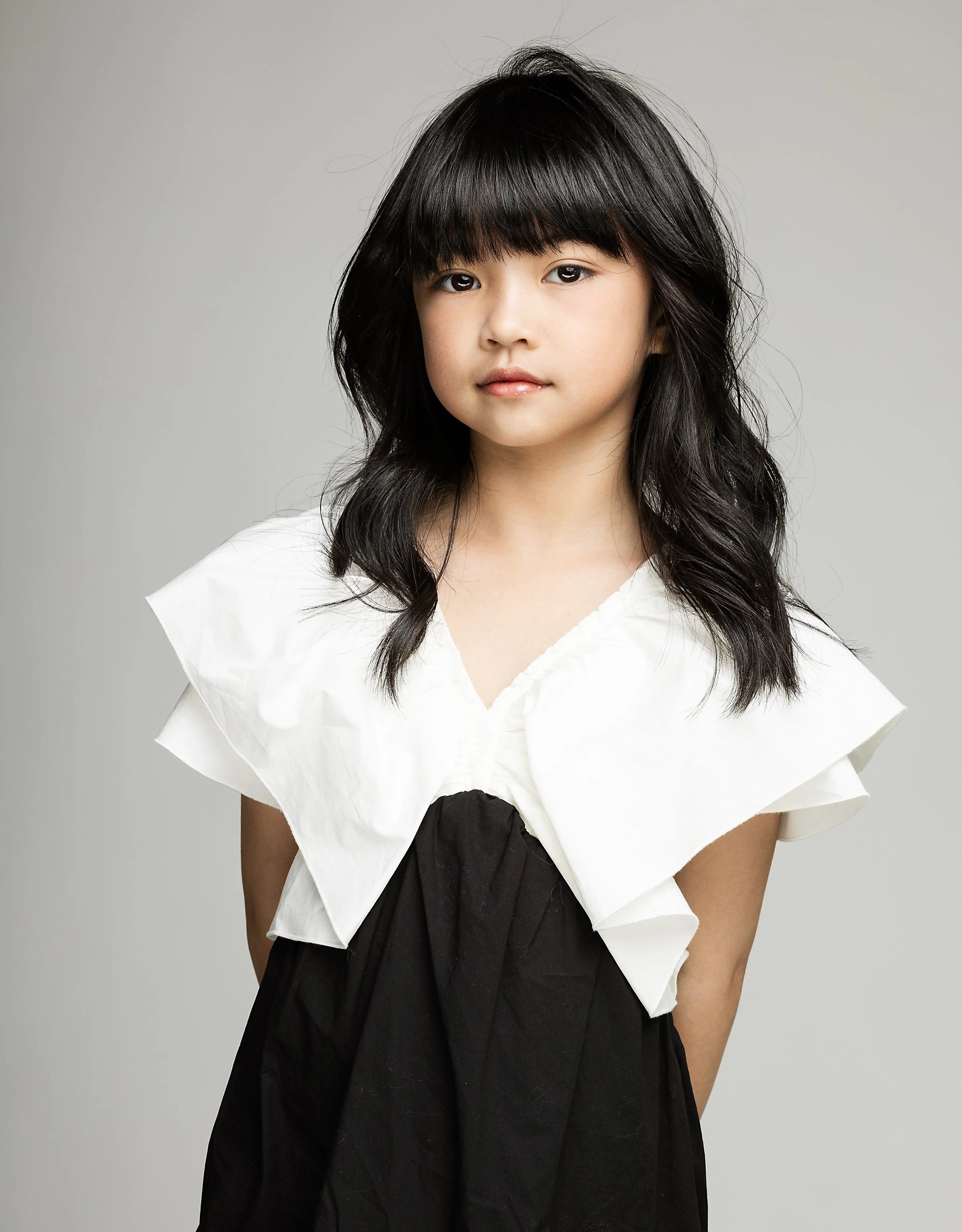 A young girl with long black hair and bangs, wearing a white top with ruffled sleeves and a black skirt, standing against a plain gray background.