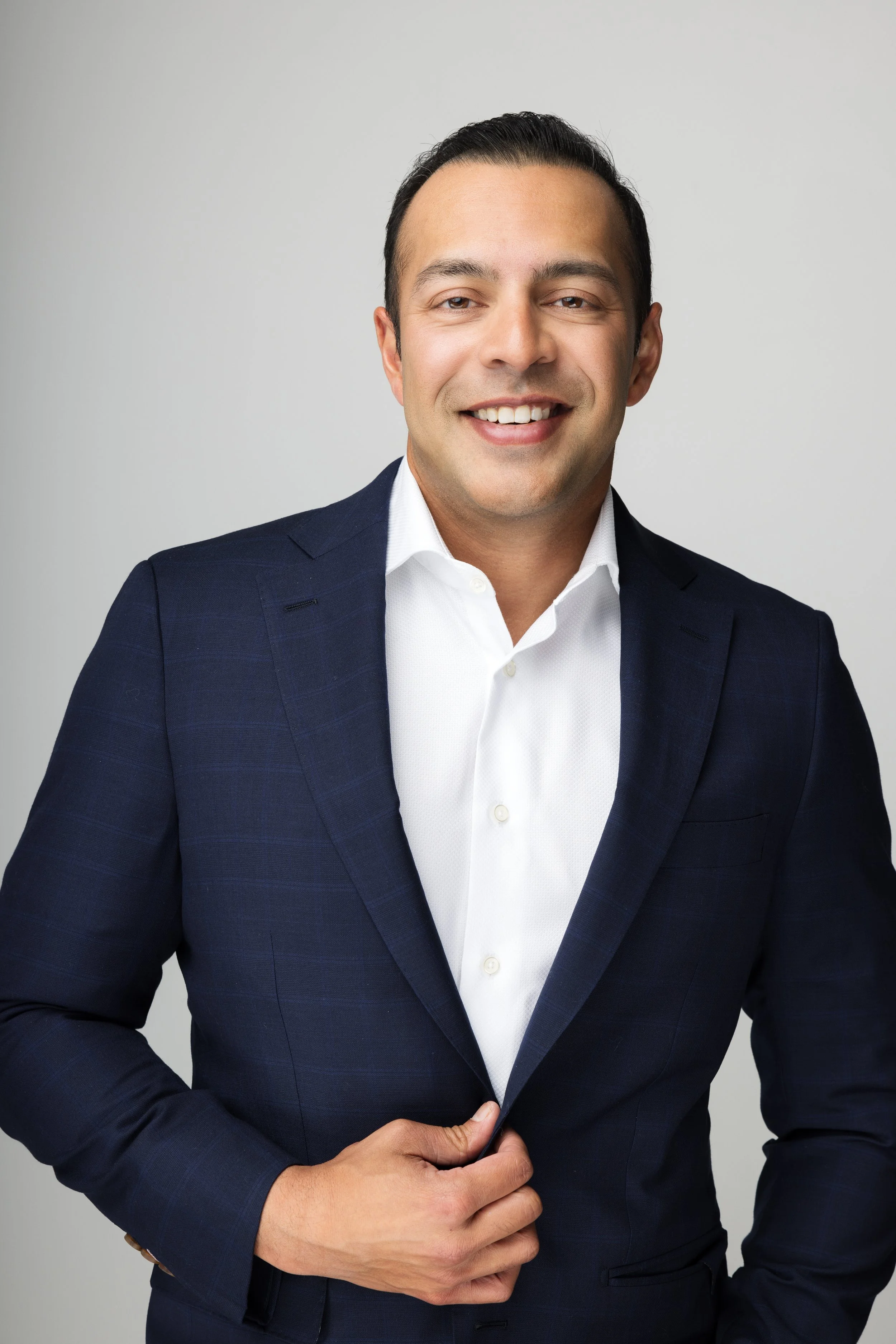 A man in a navy blue suit with a white shirt, smiling and adjusting his jacket with his left hand, standing against a plain light gray background.