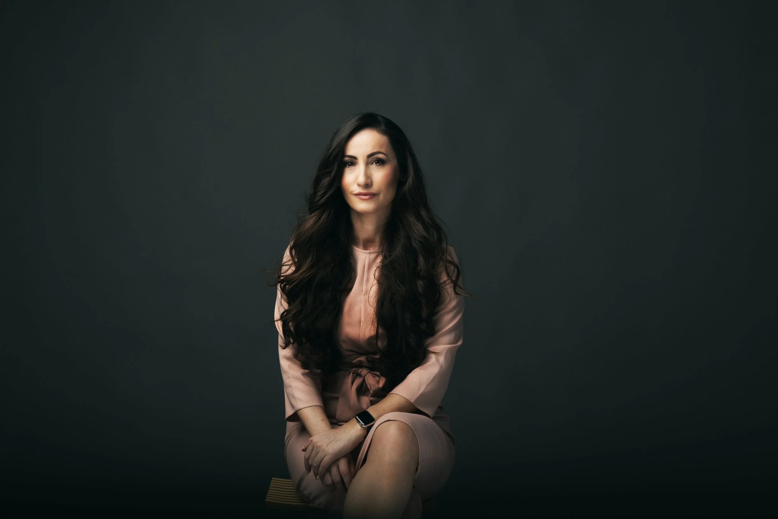 A woman with long dark hair, wearing a light pink satin dress, sitting with one knee crossed over the other, against a dark background. She has a confident expression with a slight smile.