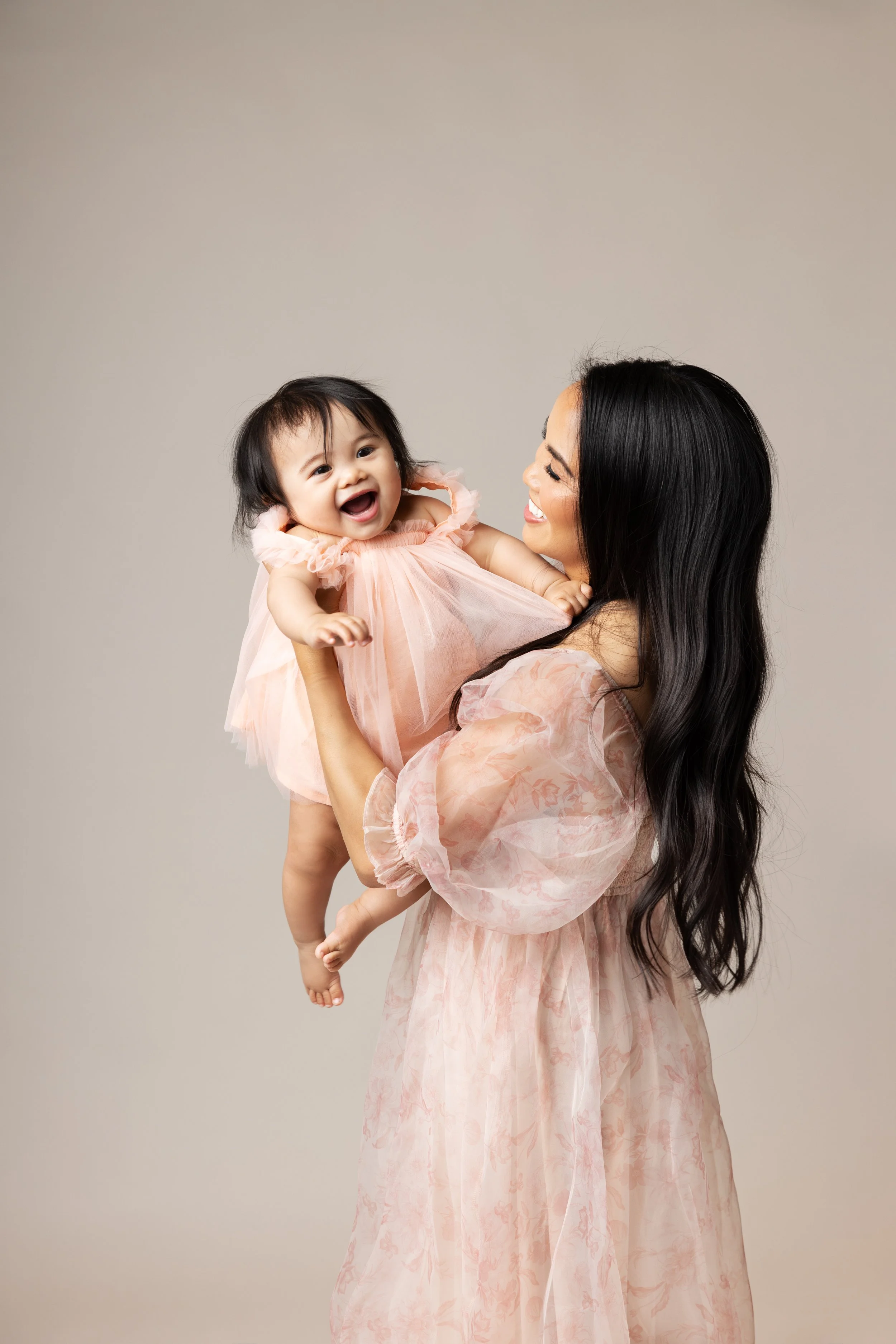 A woman with long black hair wearing a pink floral dress holding a smiling baby girl in a pink dress.