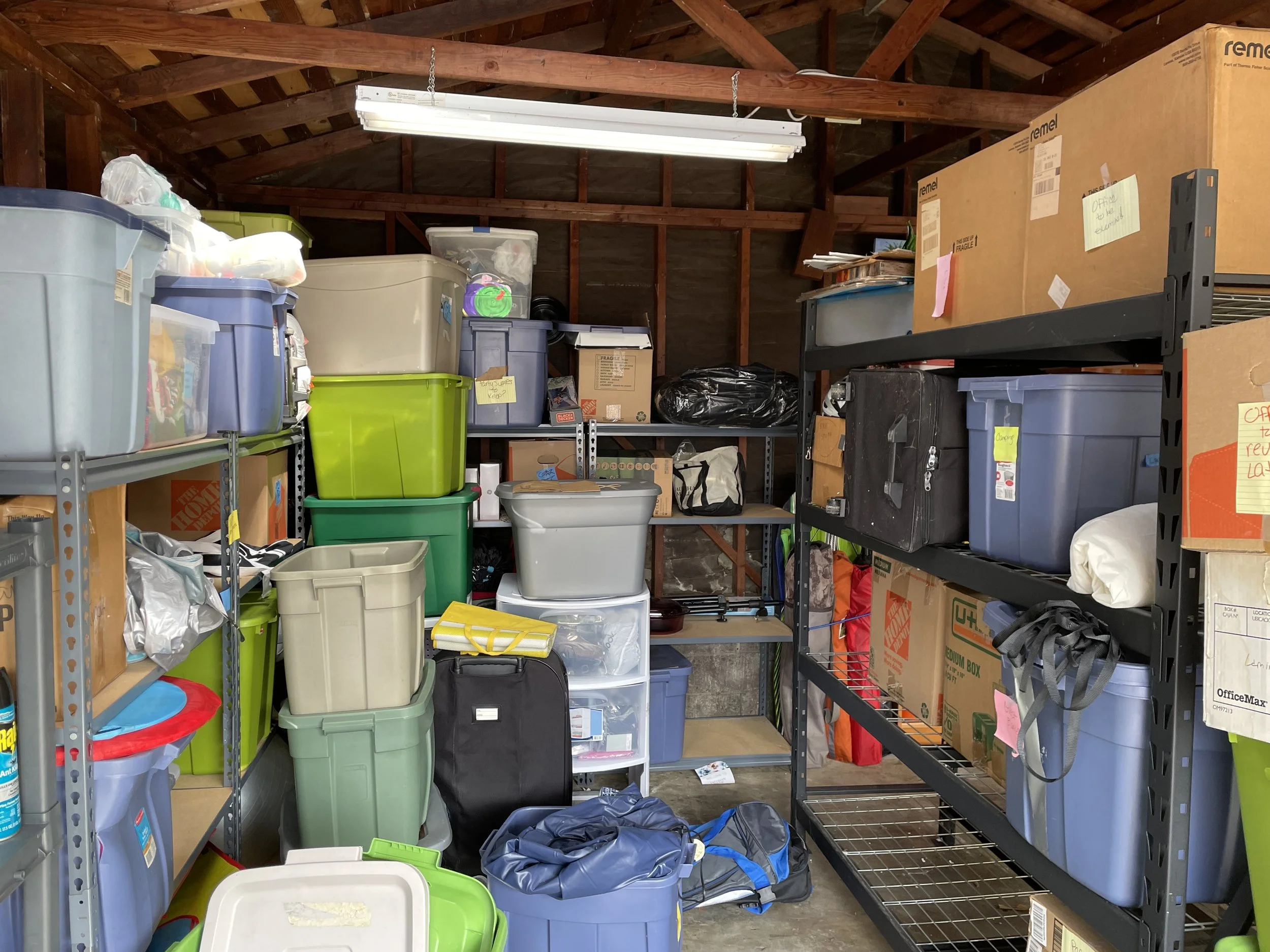 A cluttered garage with metal shelving filled with plastic storage bins, cardboard boxes, and bags, under a wooden ceiling with fluorescent lighting.