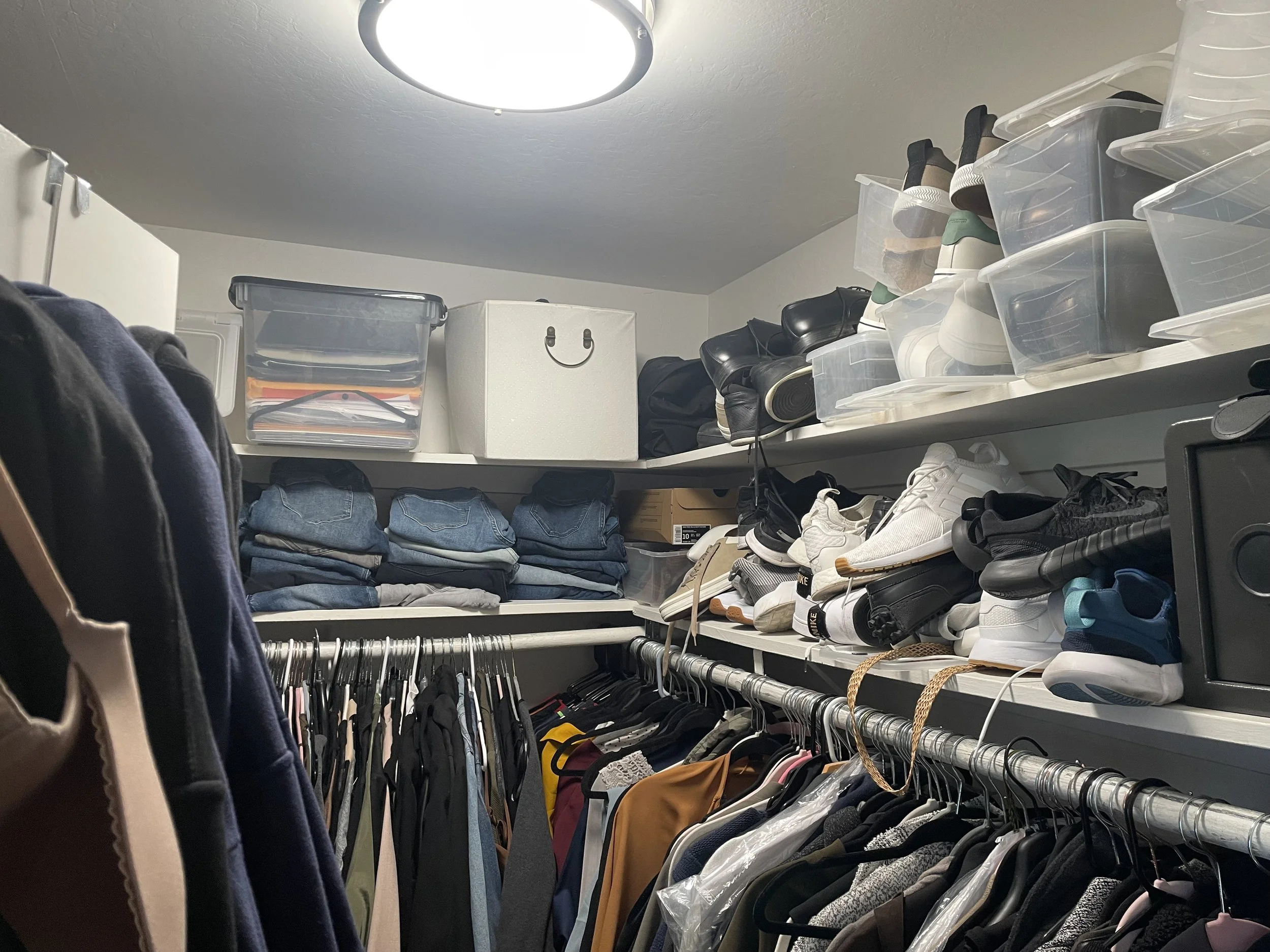 A walk-in closet with shelves of folded jeans, shoe boxes, and various sneakers, along with hanging clothes on a metal rod.