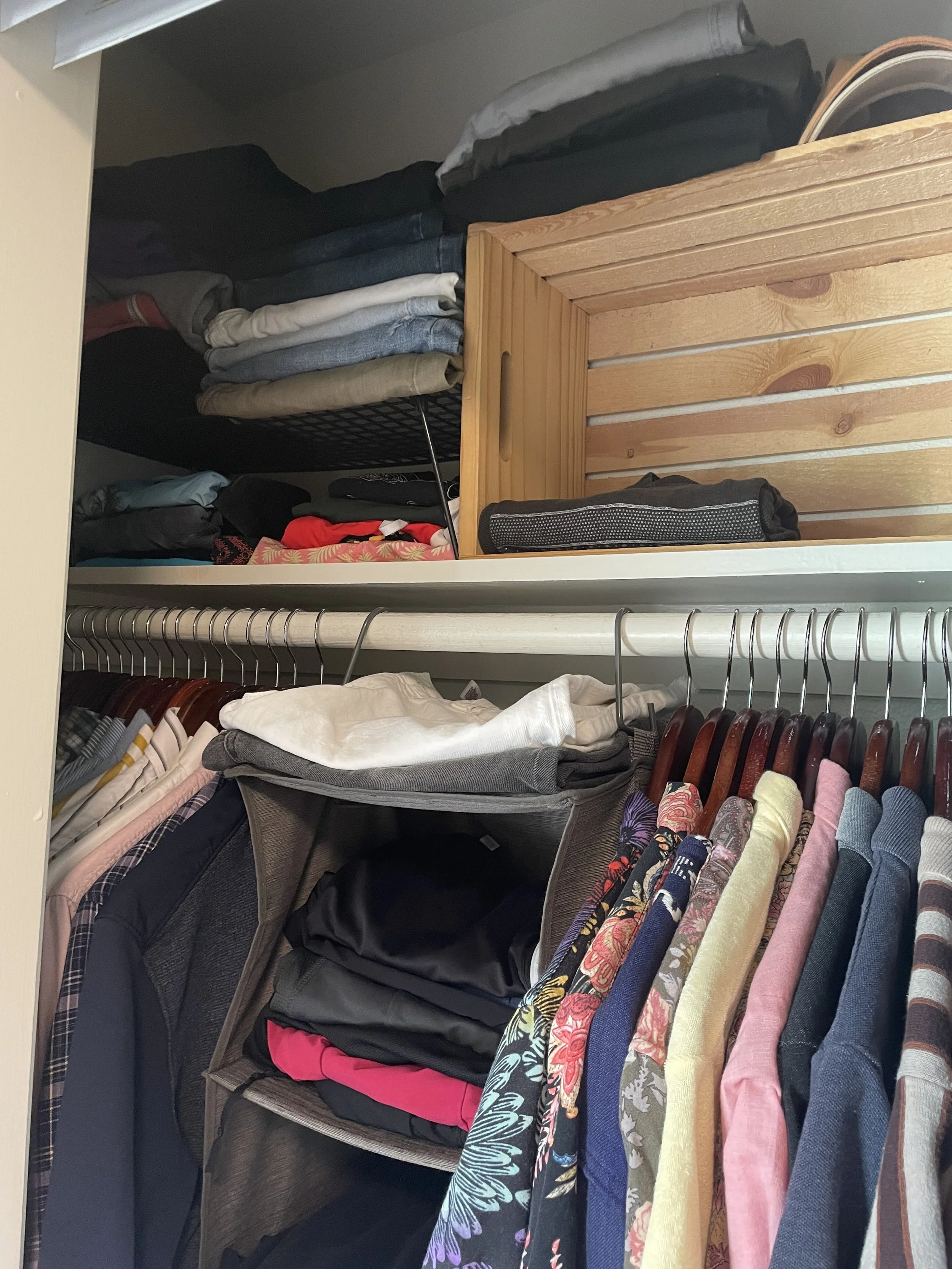 Clothes organized in a closet with a shelf on top, including folded shirts, pants, and jackets, and a hanging rod with various shirts on hangers.