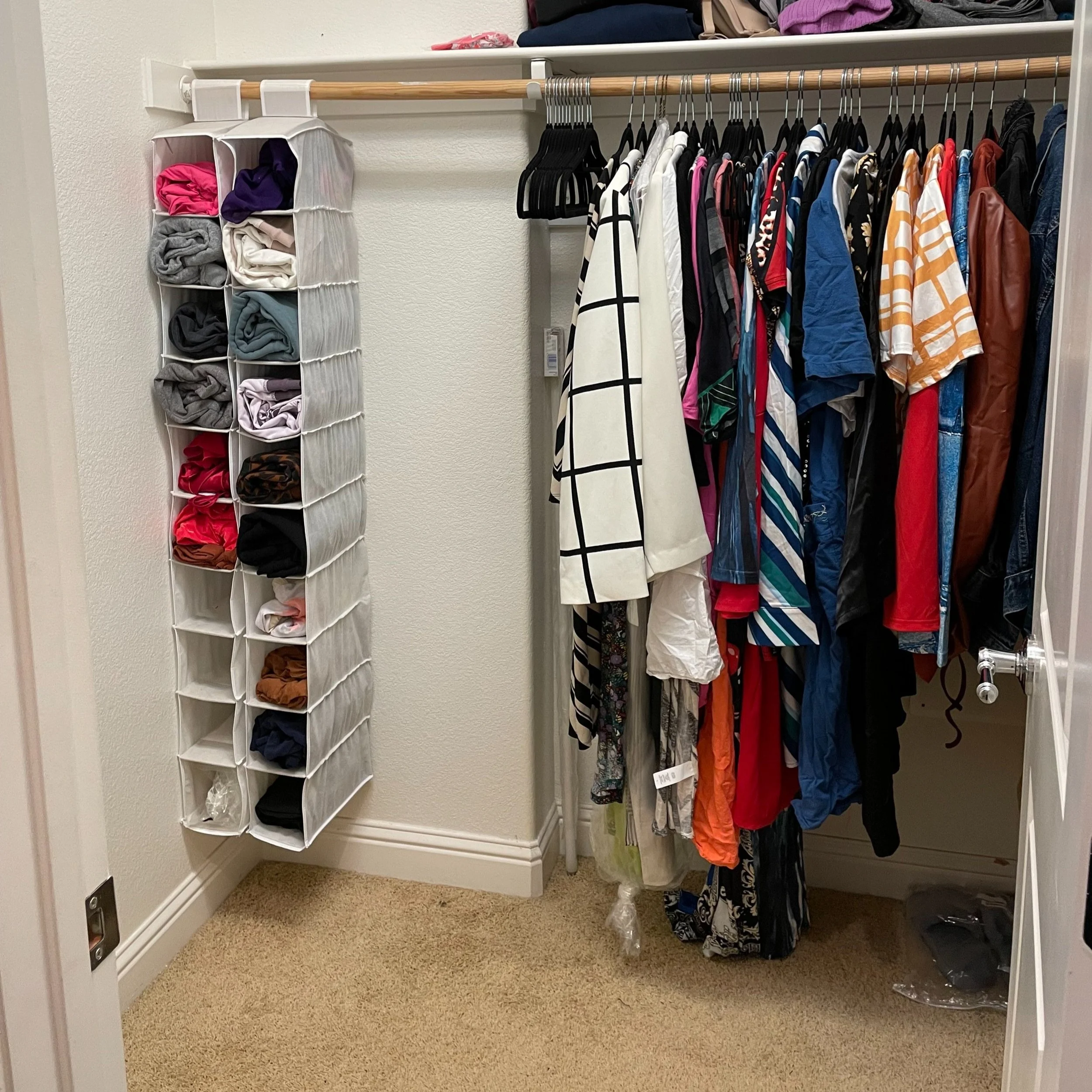 A closet with hanging clothes, including shirts, jackets, and dresses, and a hanging organizer with folded clothing items.