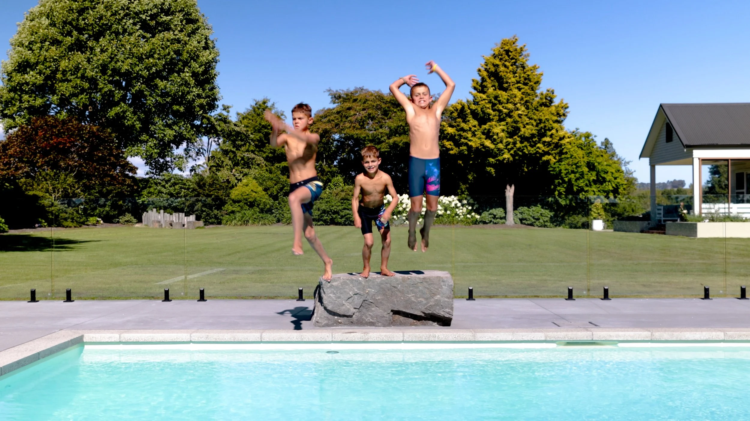 Three boys jump off a boulder beside a swimming pool