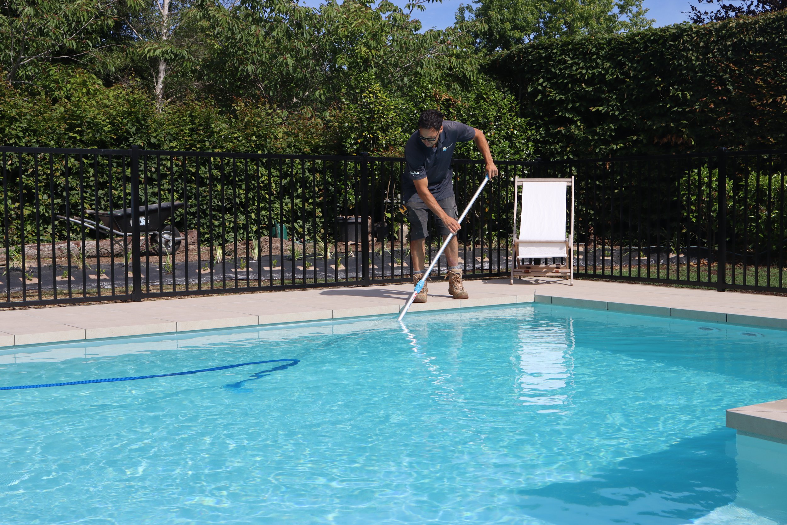 A man cleaning a swimming pool with a pool skimmer pole, surrounded by a black fence and greenery, with a white lounge chair and a wheelbarrow nearby.