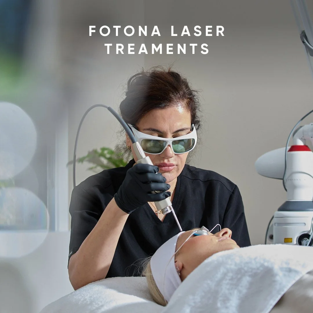 A woman wearing protective goggles and black gloves performs a laser treatment on a patient lying on a medical bed, with medical equipment nearby, in a clinical setting.
