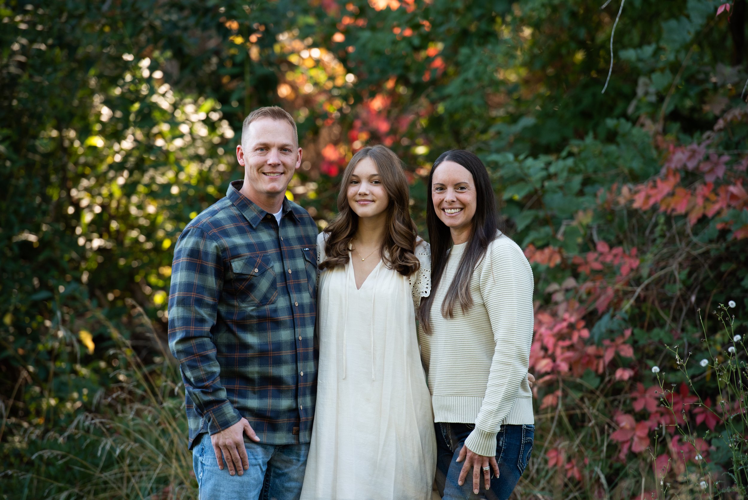 A family of three standing outdoors among green and red foliage during fall. A man, woman, and young girl are smiling at the camera. The man has short hair and is wearing a plaid shirt, the girl is in a white dress with curly brown hair, and the woman has long dark hair and is dressed in a beige sweater.