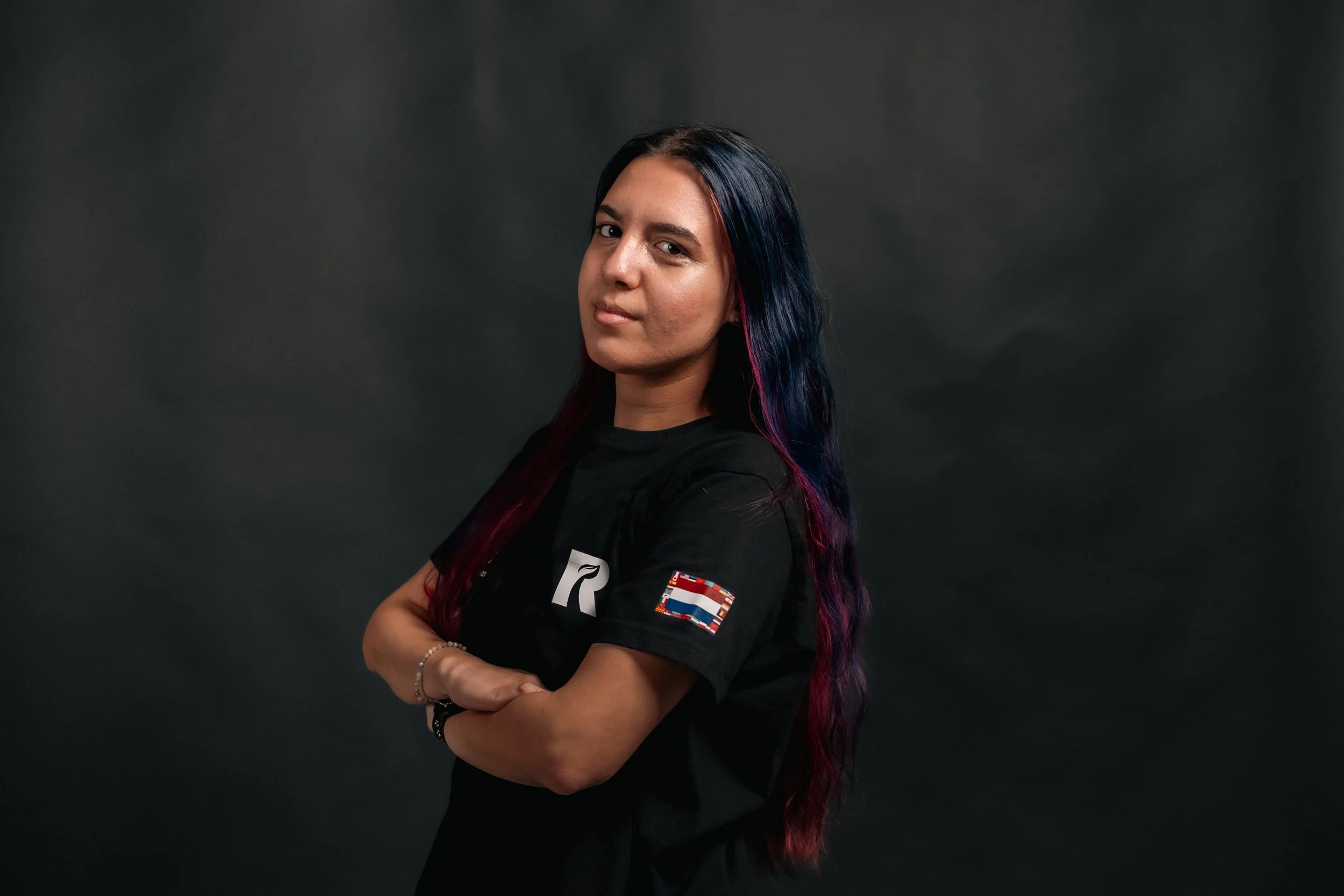 Portrait of a young woman with long dark hair, wearing a black t-shirt with a Dutch flag patch on the sleeve, standing against a dark background.