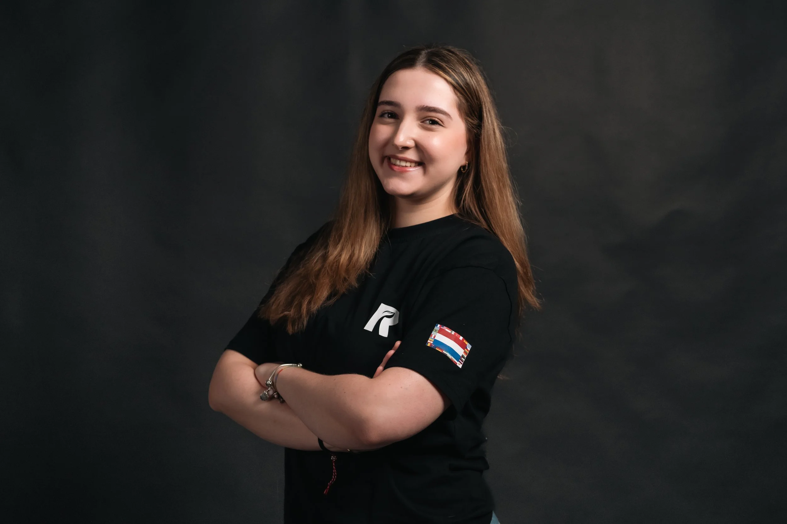 A young woman with long, wavy blonde hair and glasses, wearing a black t-shirt with logos, standing against a dark background.