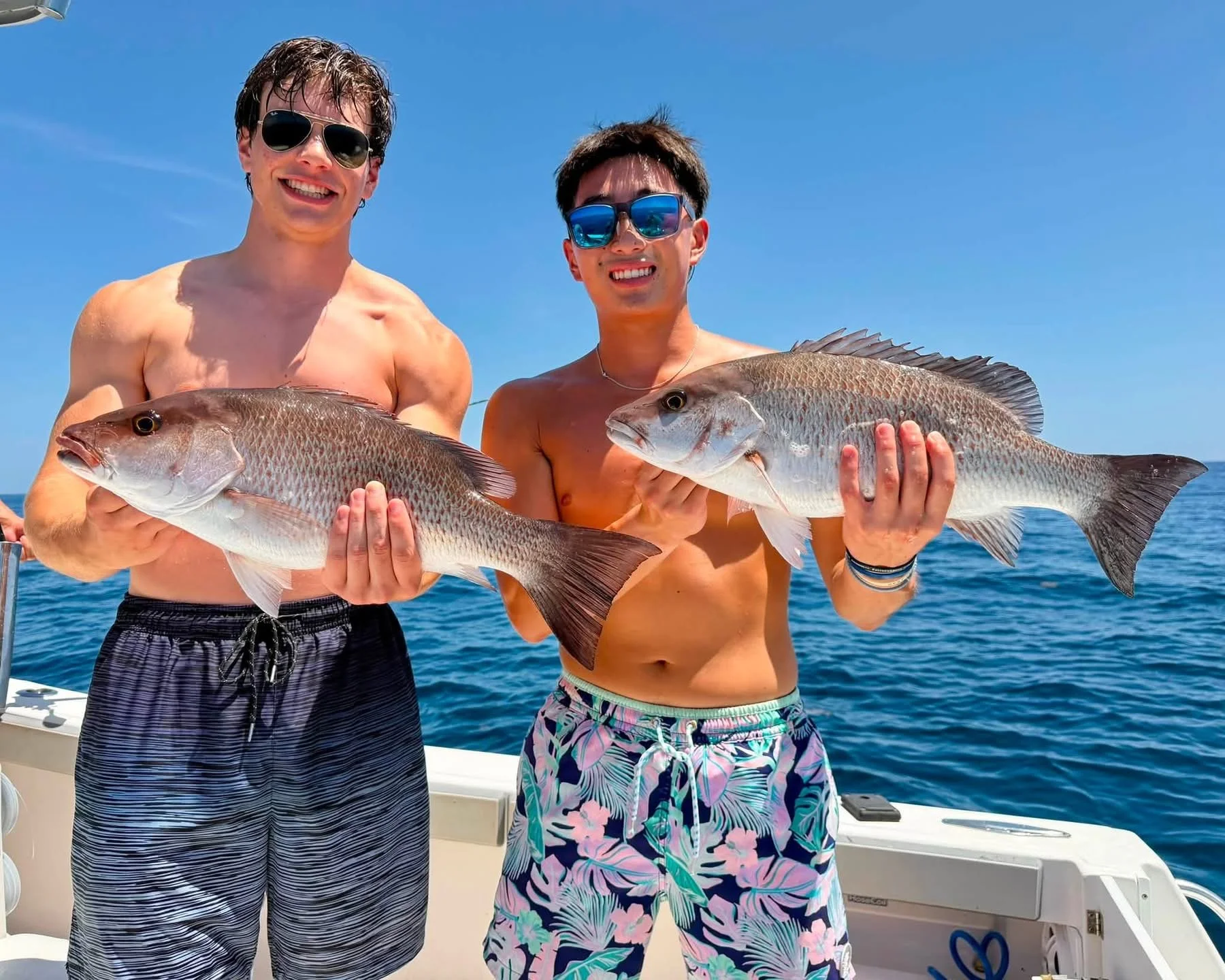 Man in sunglasses and neon green shirt holding a large fish on a boat with fishing rods in the background, out at sea on a clear day.