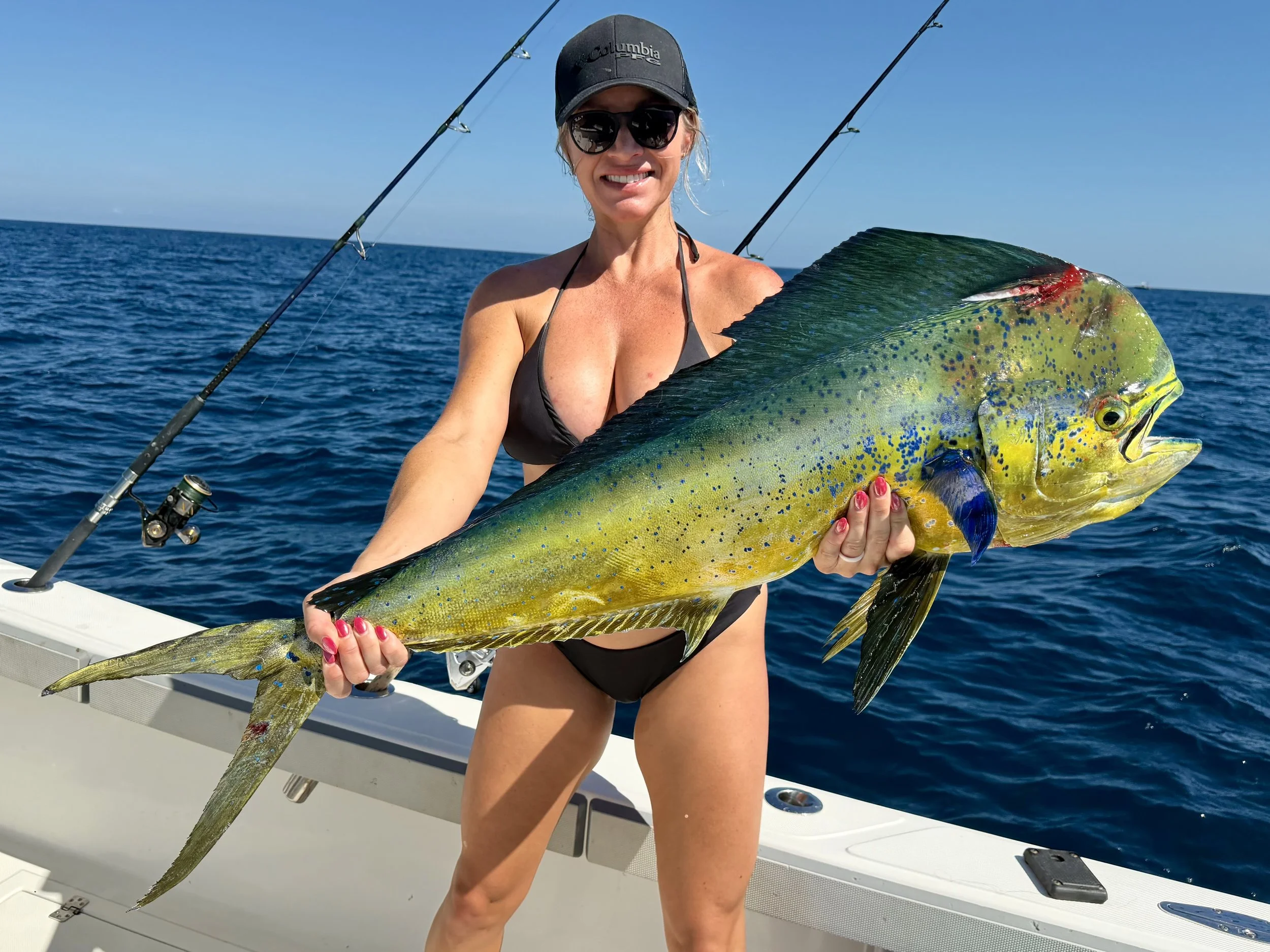 Three women on a boat holding a large fish, smiling, with fishing rods behind them and the ocean surrounding the boat.