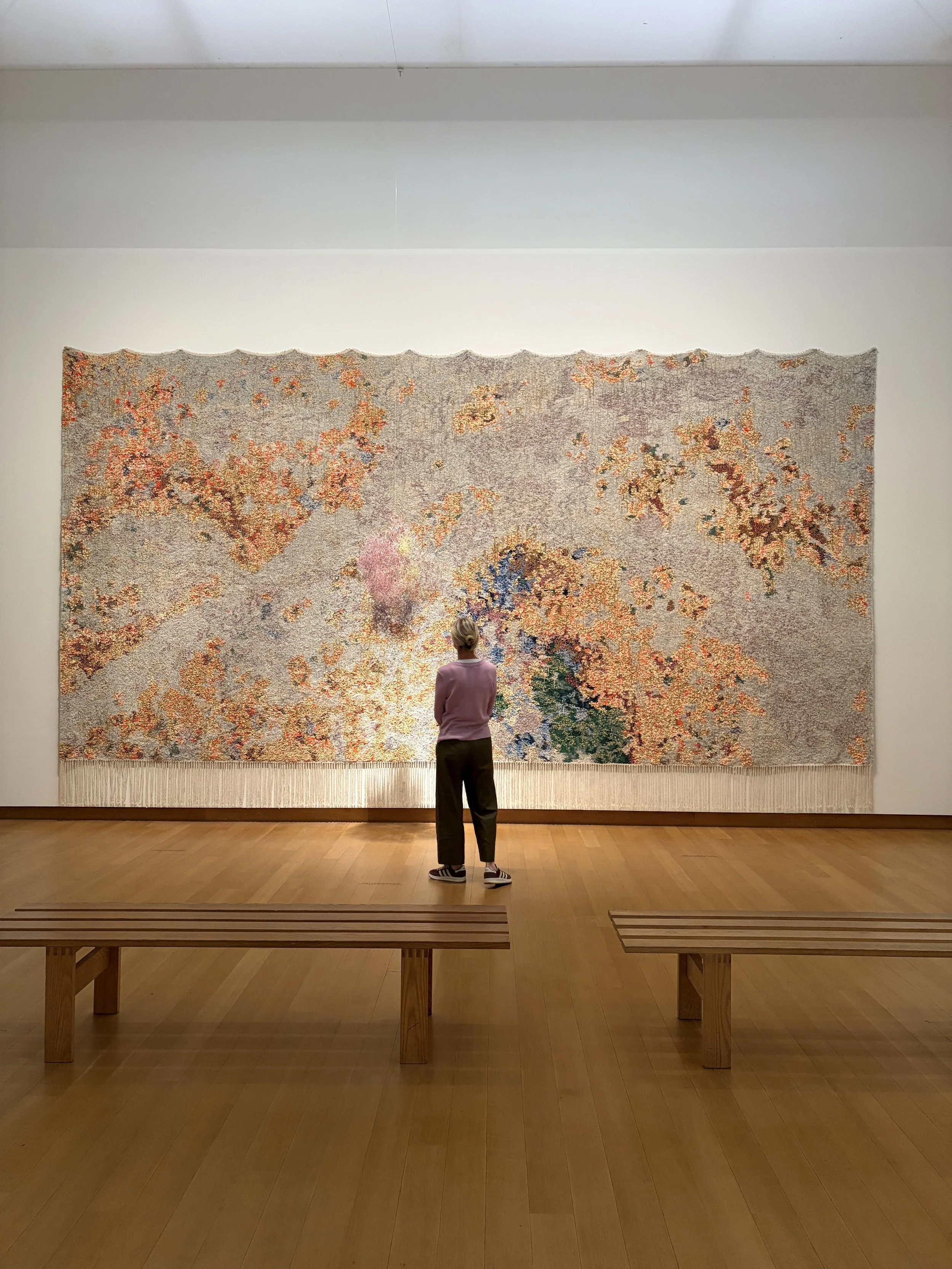 A person with blonde hair, wearing a pink sweatshirt and striped sneakers, stands facing a large abstract textured artwork in a gallery. The artwork features small, scattered, colorful patches on a neutral background, resembling a map or mosaic. The gallery has wooden flooring and a display bench in the foreground.