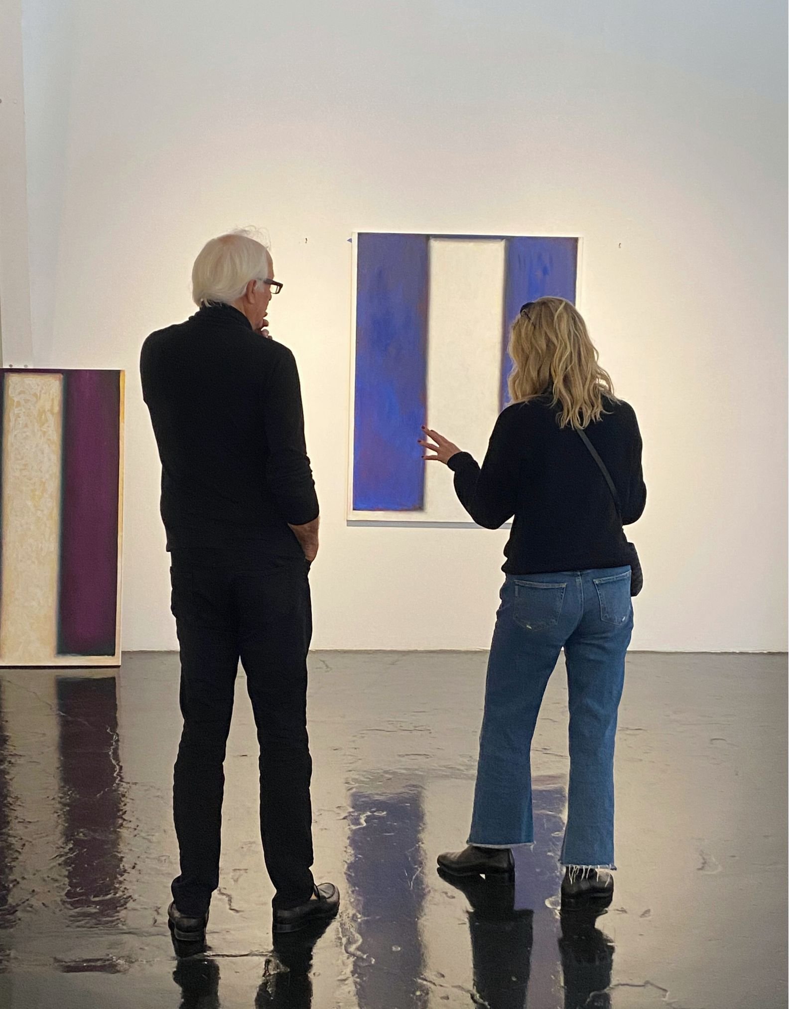 Two people, a man with white hair and glasses and a woman with blonde hair, are standing in front of a white wall with artwork. They are looking at a large painting consisting of two vertical blue rectangles. They are standing in a space with a shiny, reflective floor.