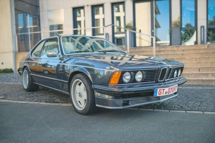 Ein blauer, alter BMW E24 6er mit deutschen Nummernschildern, geparkt vor einer modernen Gebäudefassade.