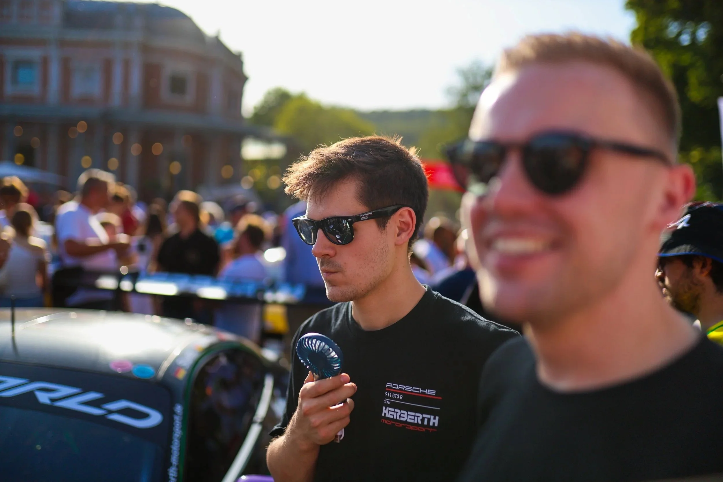Zwei junge Männer tragen Sonnenbrillen bei einem Motorsport-Event, im Hintergrund eine Menschenmenge und ein historisches Gebäude, sonniges Wetter.