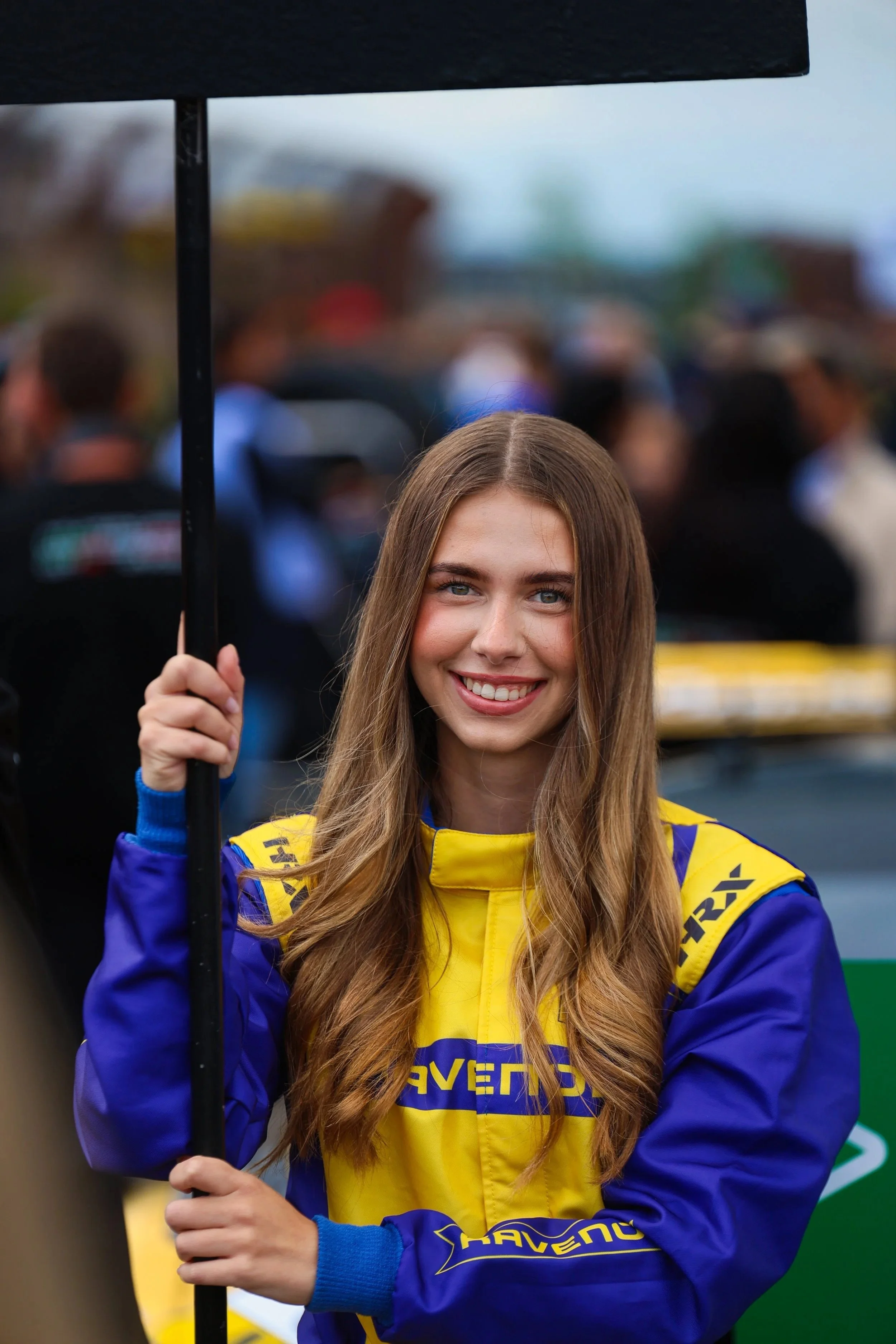 Eine junge Frau trägt einen blauen und gelben Rennfahreranzug und hält einen schwarzen Regenschirm bei einem Motorsportevent.