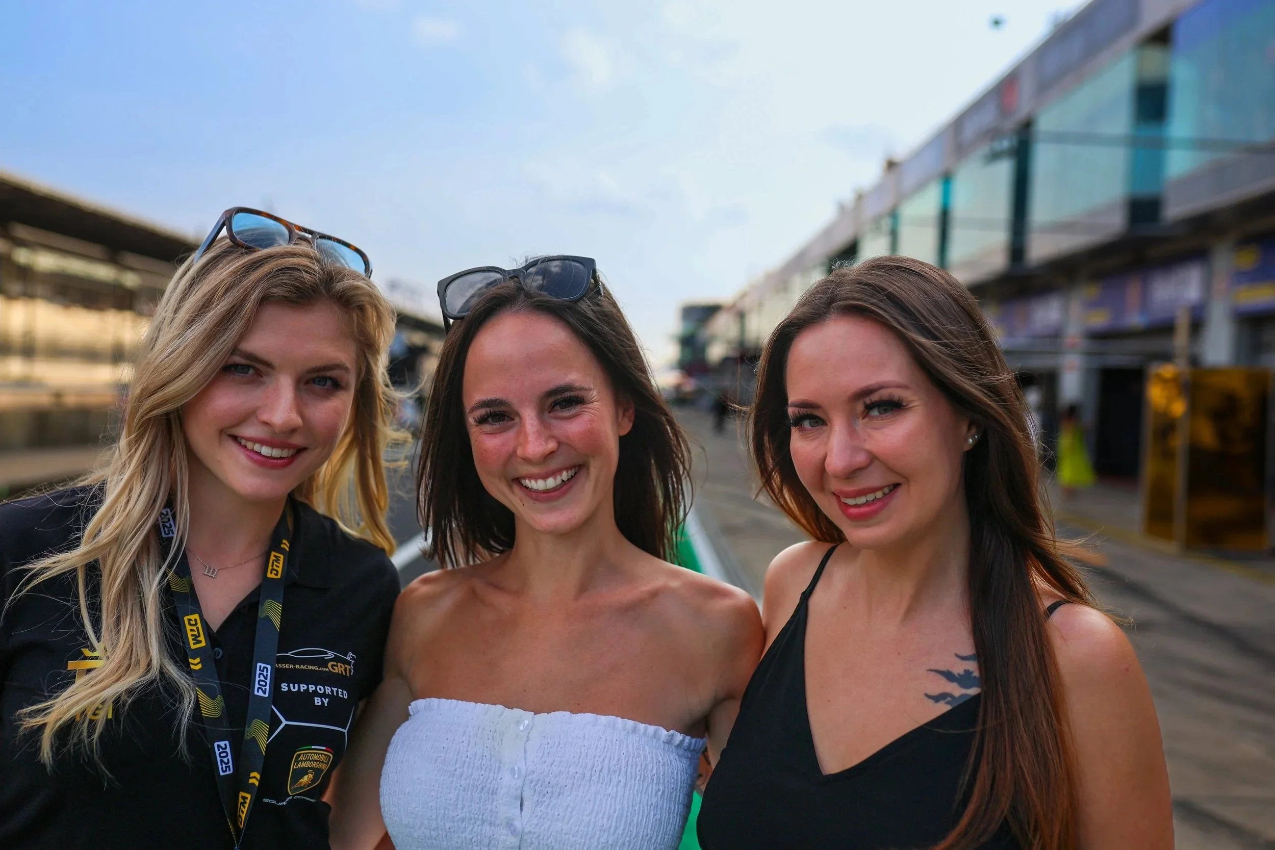 Drei junge Frauen stehen nebeneinander vor einer Rennstrecke, lächeln und posieren für das Foto.