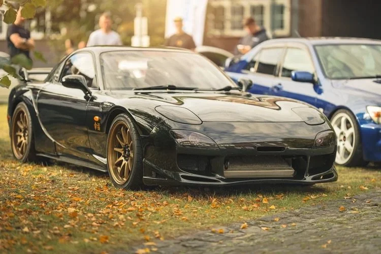Schwarzer Sportwagen mit goldenen Felgen auf einem Rasen bei einem Autoevent, Herbstblätter am Boden, im Hintergrund Menschen und andere Autos.