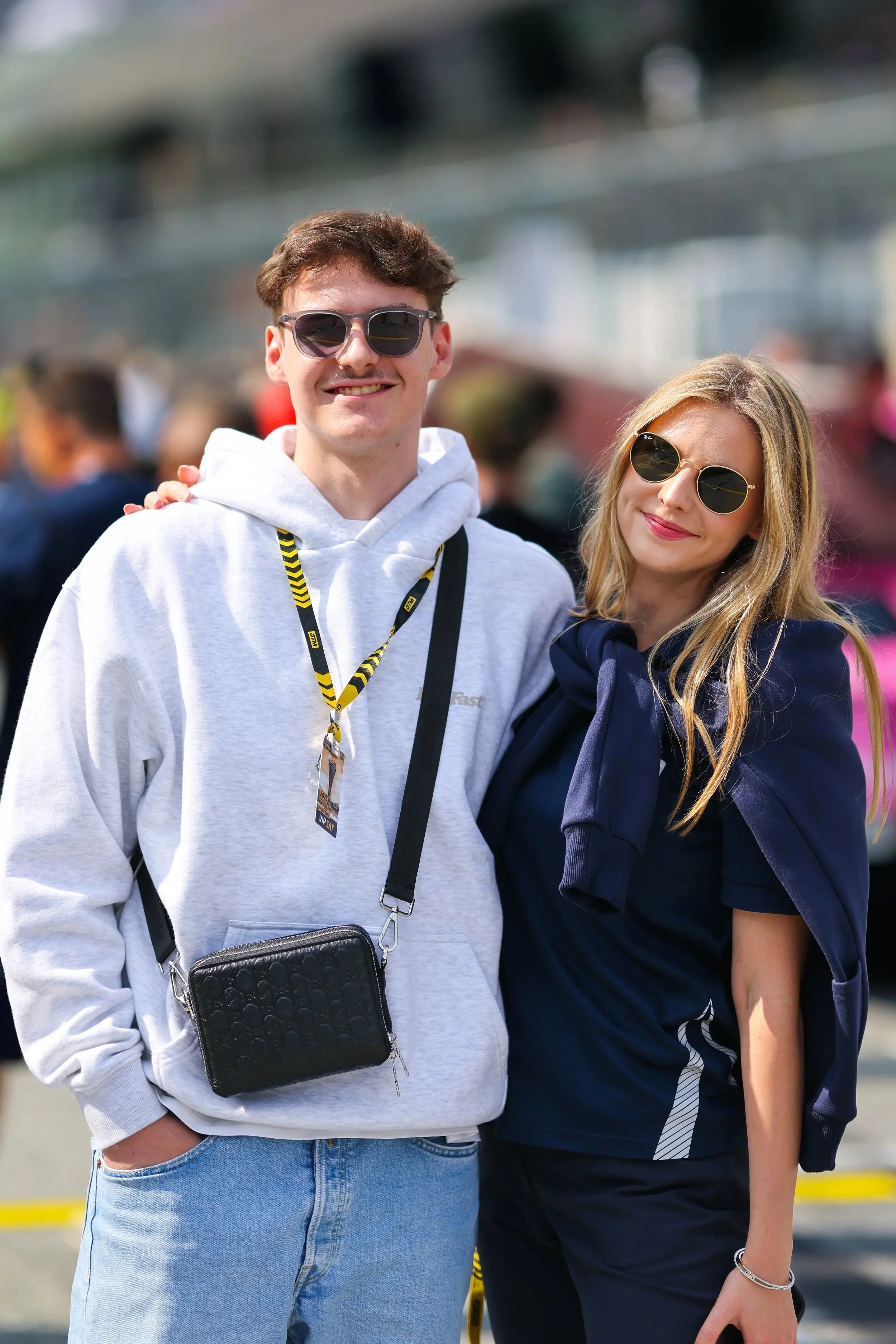 Zwei junge Menschen, ein Mann und eine Frau, stehen im Freien, lächeln und tragen Sonnenbrillen. Der Mann trägt ein weißes Hoodie mit schwarzer Tasche, die Frau trägt ein dunkelblaues Oberteil mit einer Jacke um die Schultern.