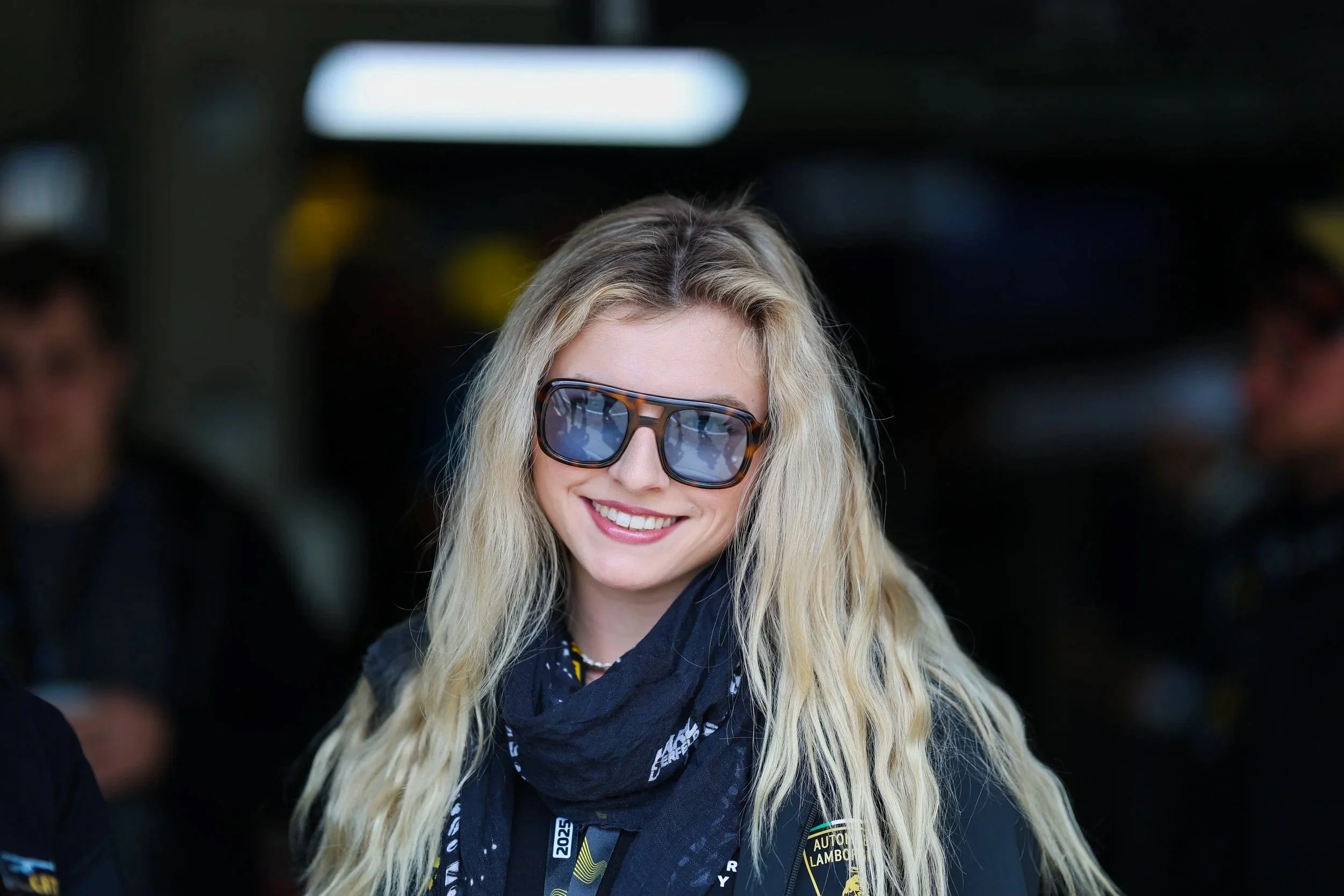 Junge Frau mit langen blonden Haaren, Sonnenbrille, lachend, in einem schwarzen Motorsport-Outfit mit Lamborghini Logo, im Hintergrund unscharfe Menschen.