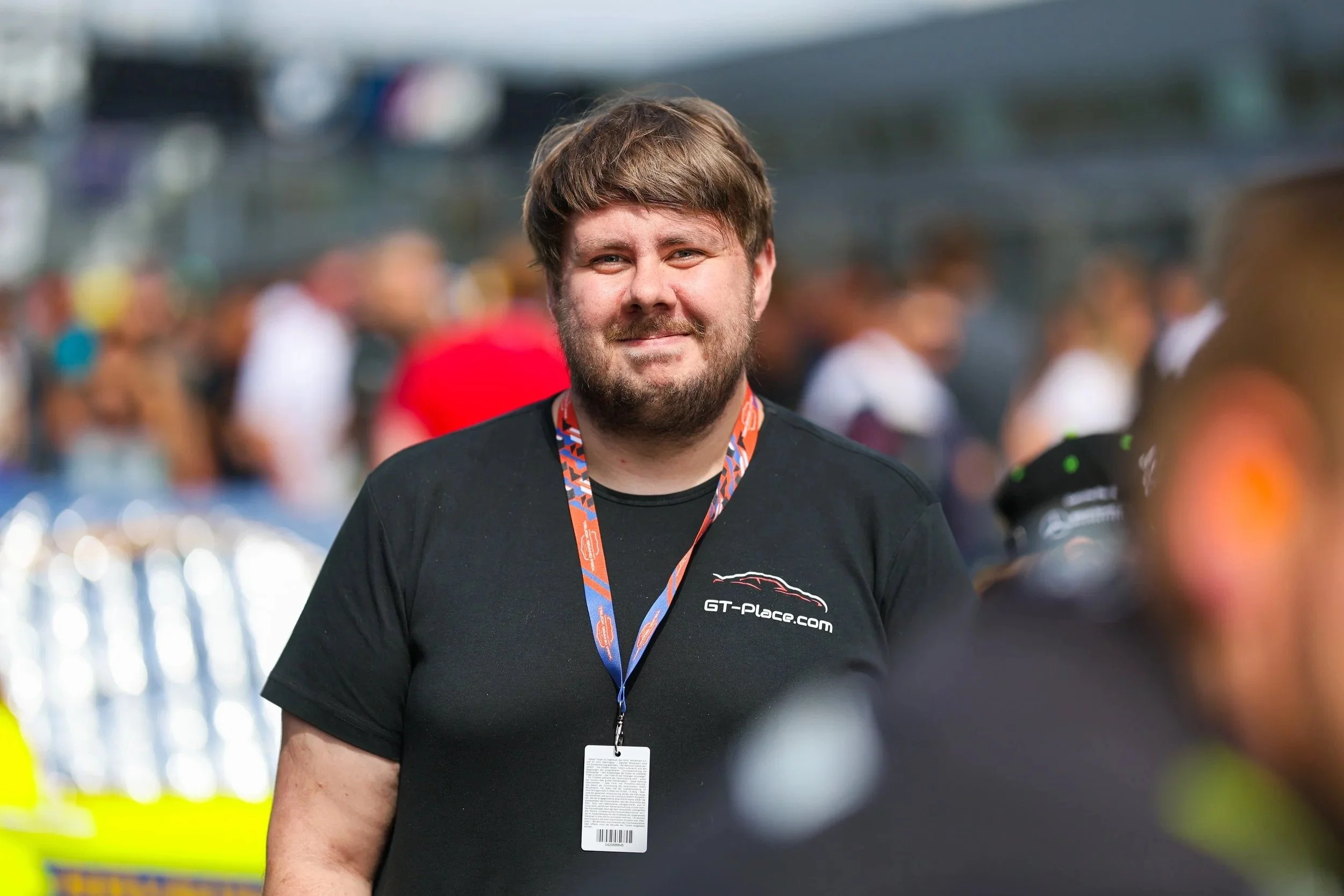 Ein Mann mit braunem Haar und Vollbart, trägt ein schwarzes T-Shirt mit dem Logo 'GT-Place.com' und eine orange-blau Gemusterte Lanyard, steht in einer Menschenmenge bei einem Event im Freien.