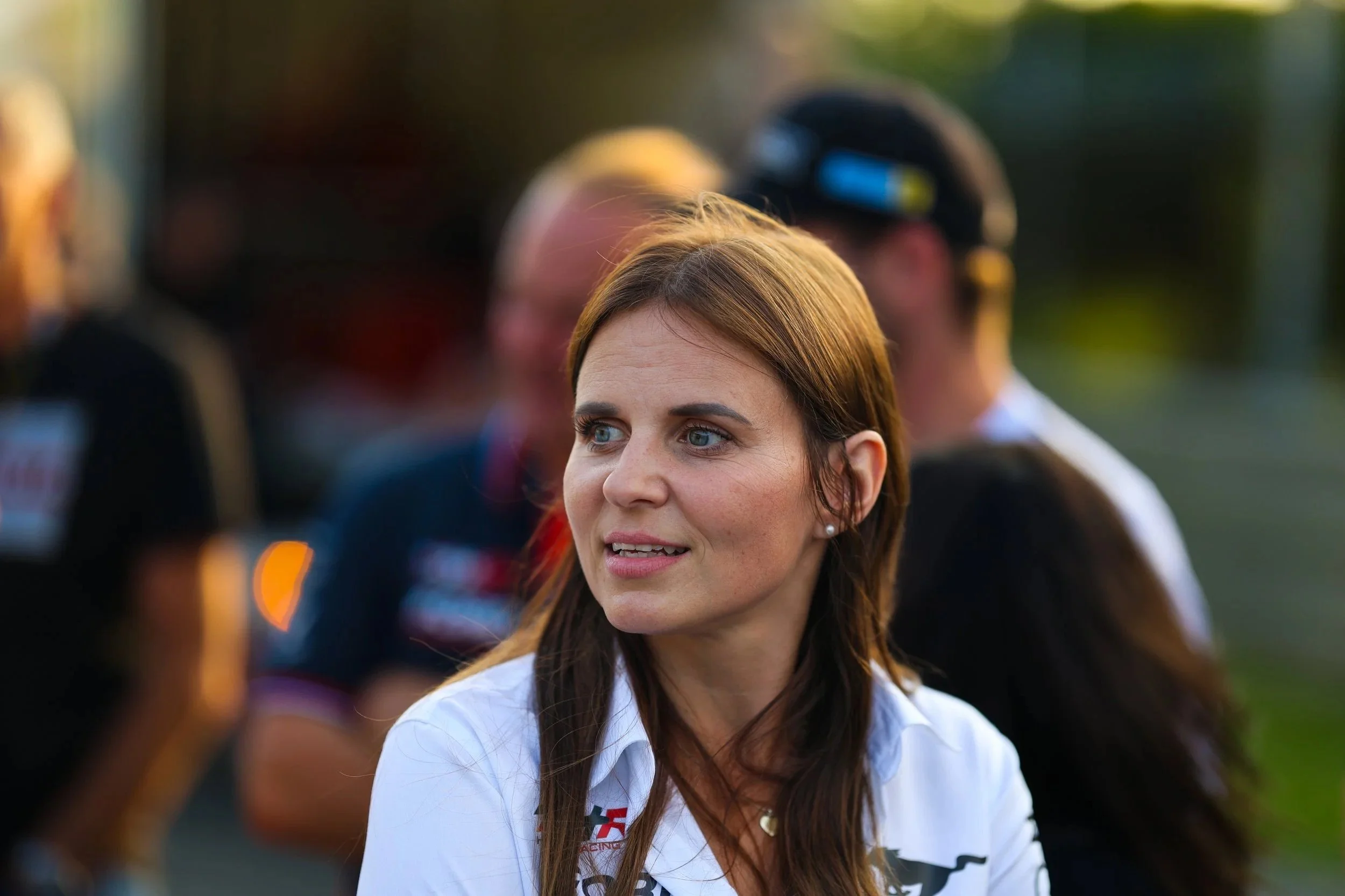 Junge Frau im weißen T-Shirt, im Hintergrund mehrere Menschen, wahrscheinlich bei einem Motorsport-Event, mit einem Fokus auf die Frau, sonniges Wetter, Naturhintergrund.