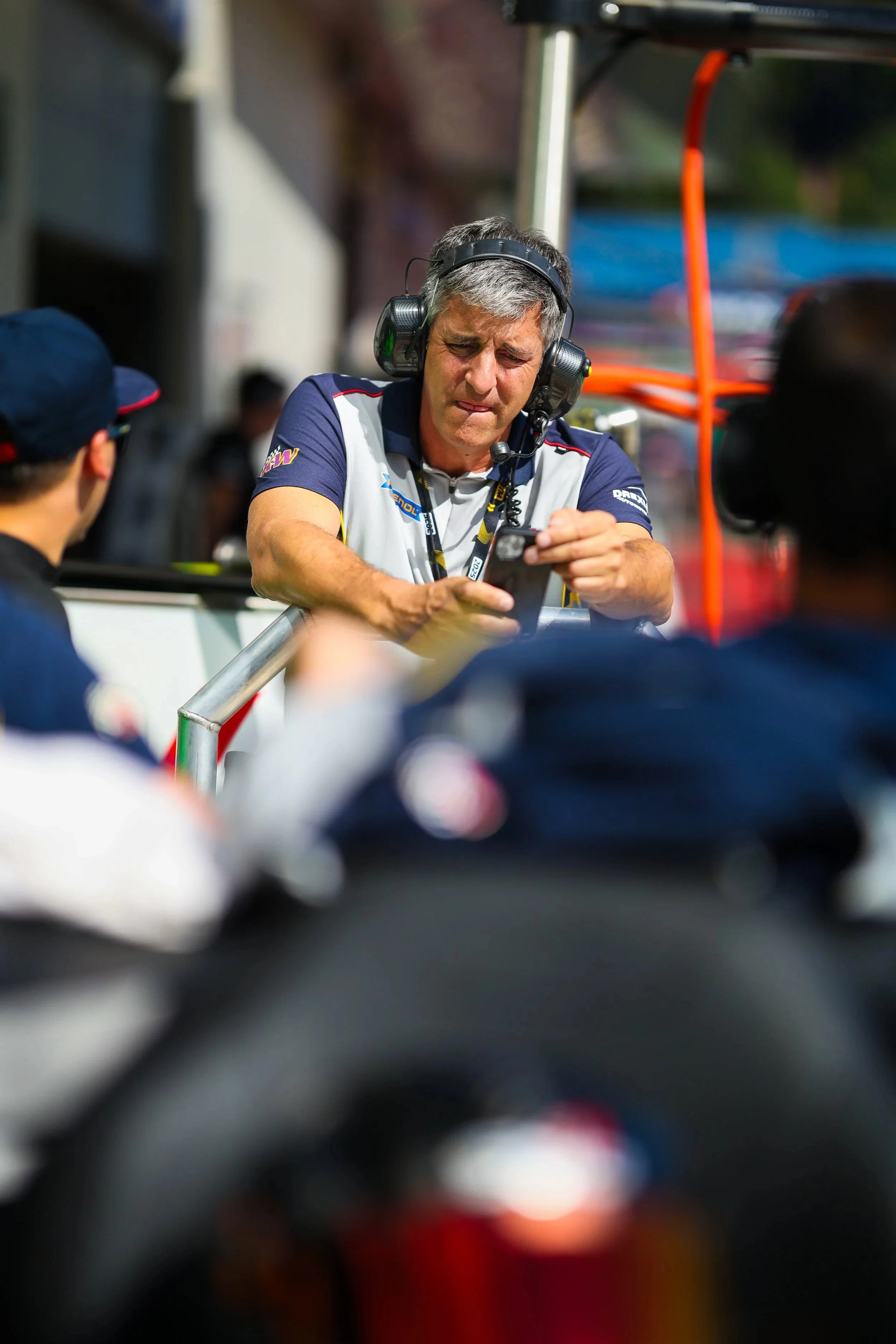 Ein Mann mit Kopfhörern und einem Team-Shirt, der auf einem Motorsport-Event mit einem Smartphone beschäftigt ist, im Hintergrund ein Fahrzeug und andere Personen.