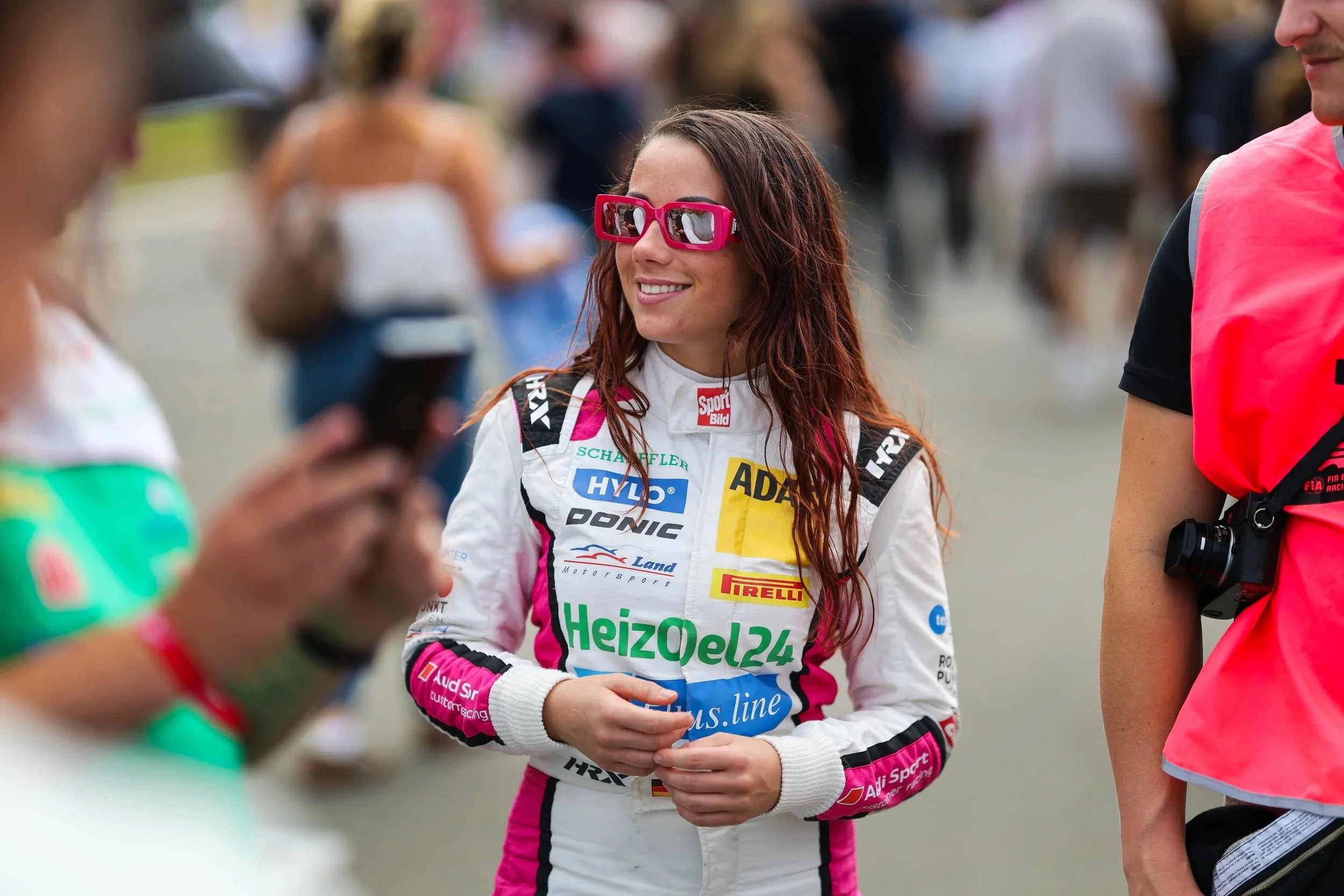 Eine junge Rennfahrerin mit roten Haaren und pinken Sonnenbrillen, trägt einen weißen Rennanzug mit verschiedenen Sponsor-Logos, steht im Gespräch mit anderen Personen bei einem Motorsport-Event.