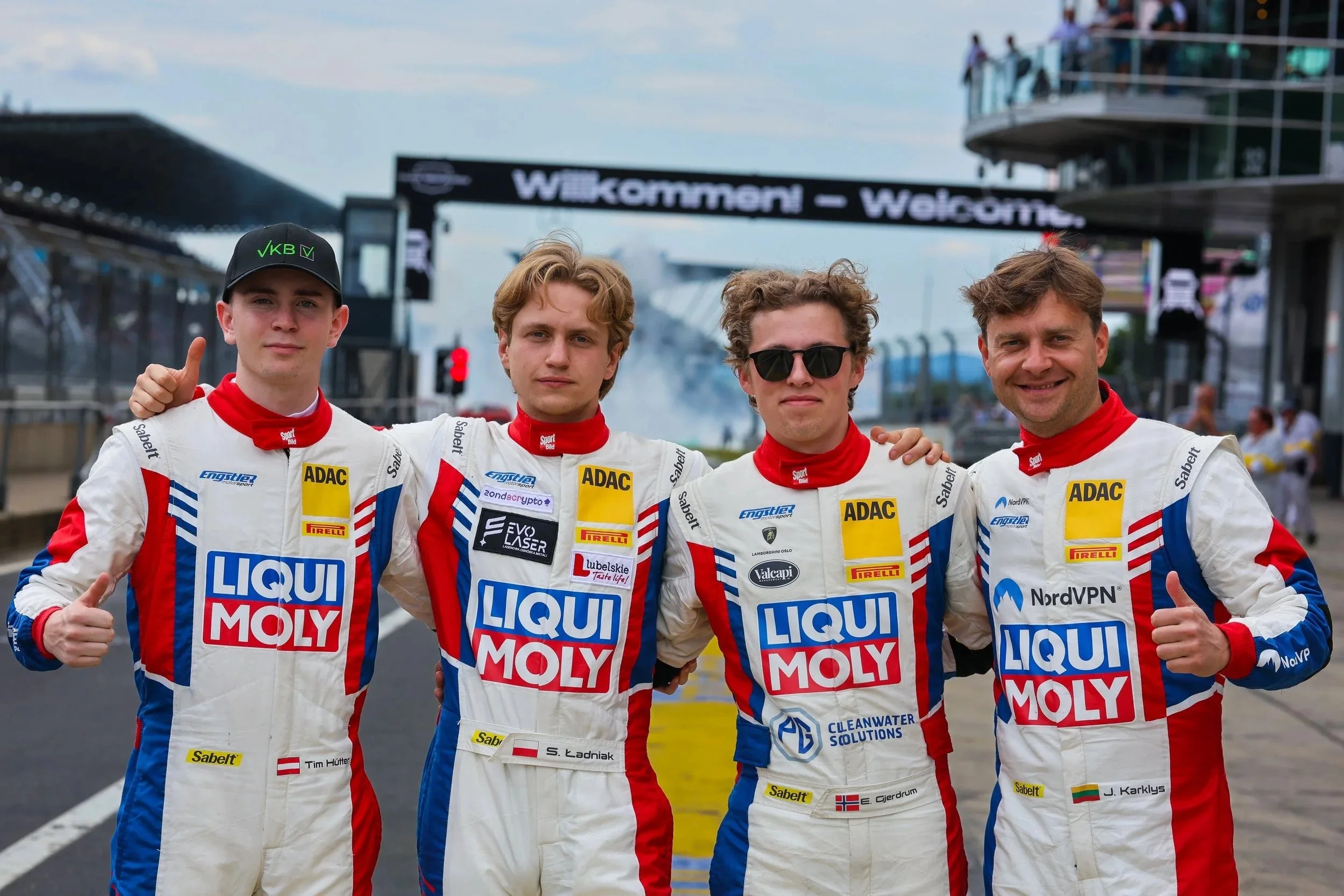 Vier Rennfahrer stehen in ihren weißen, blauen und roten Rennanzügen mit verschiedenen Sponsor Logos, sie stehen arm in arm auf dem Podium der Rennstrecke, einige sind am jubeln und posieren für die Kamera.