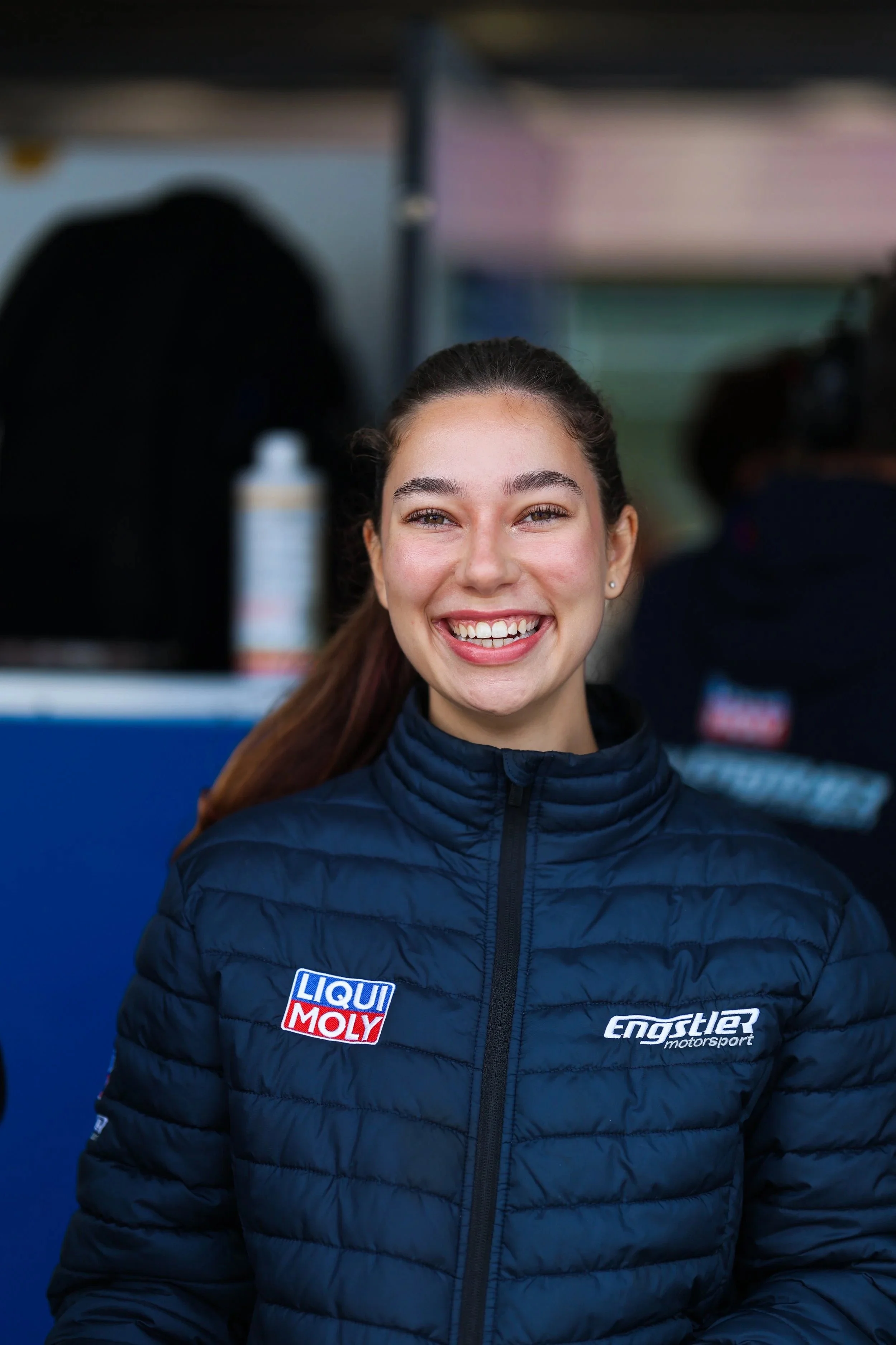 Junge Frau bei Motorsportveranstaltung, lächelnd, trägt eine blaue Jacke mit 'LIQUI MOLY' und 'Engstler Motorsport'-Aufnähern.
