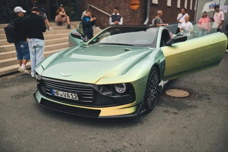 Ein silberner Aston Martin V12 Vantage am Straßenrand, umgeben von Menschen auf einer Stadtstraße.