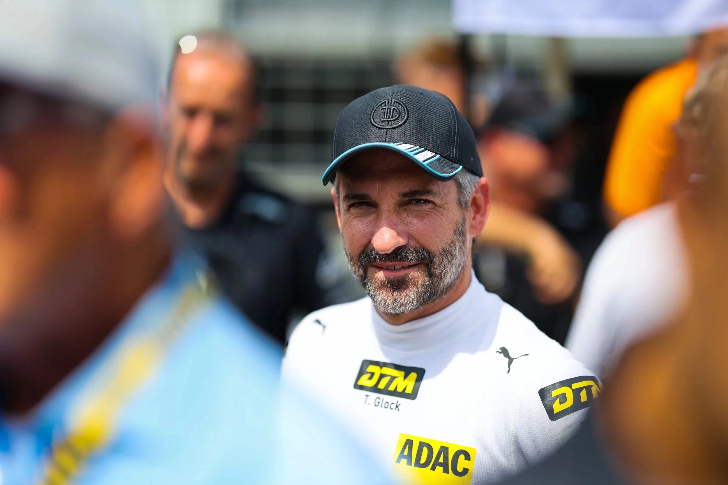 Mann mit Bart und schwarzen Hut, getragen von Sponsorenlogos, in einer Gruppe bei einem Motorsport-Event.